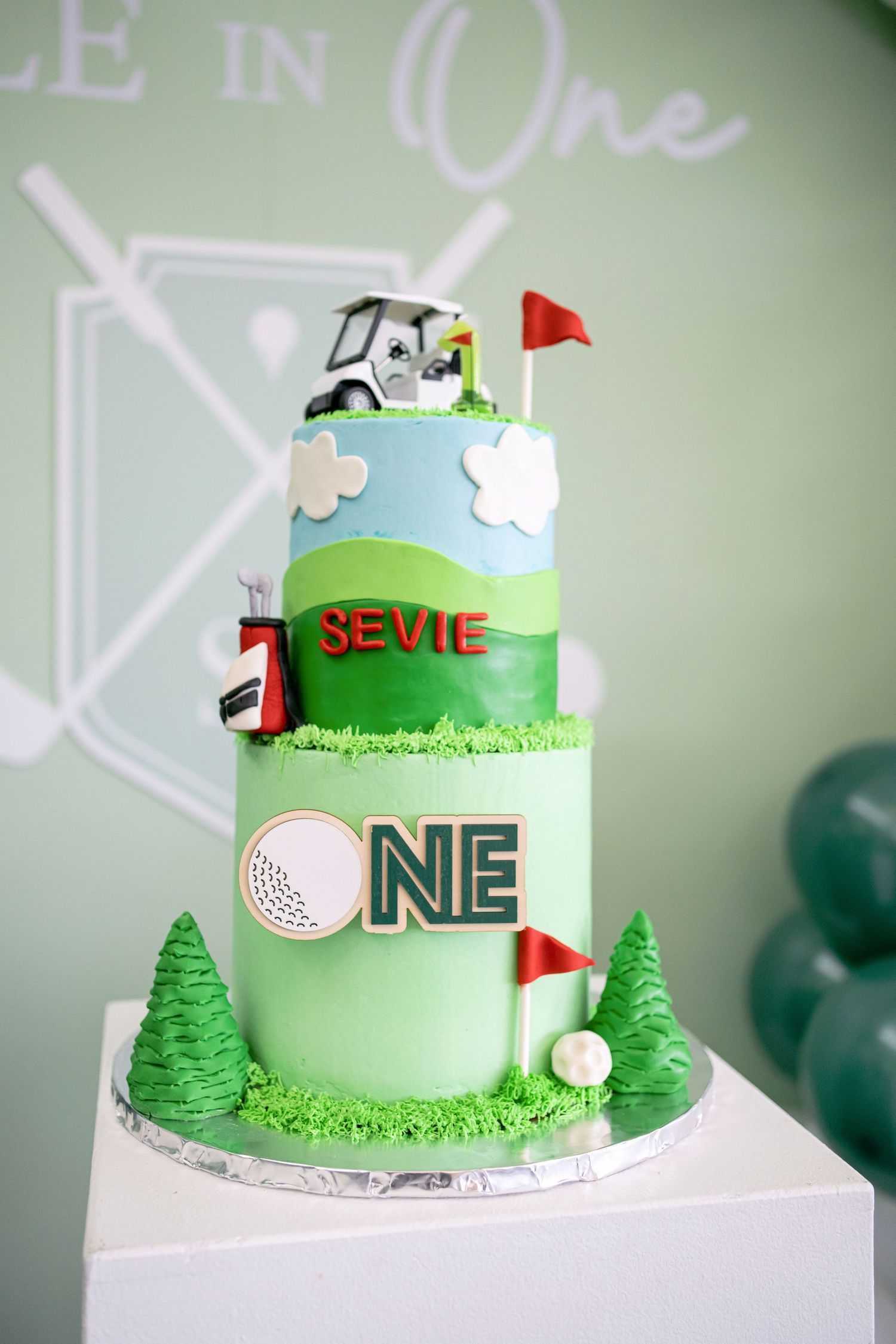A green and white golf-themed cake featuring miniature trees, flags, and golf balls on a tiered design.