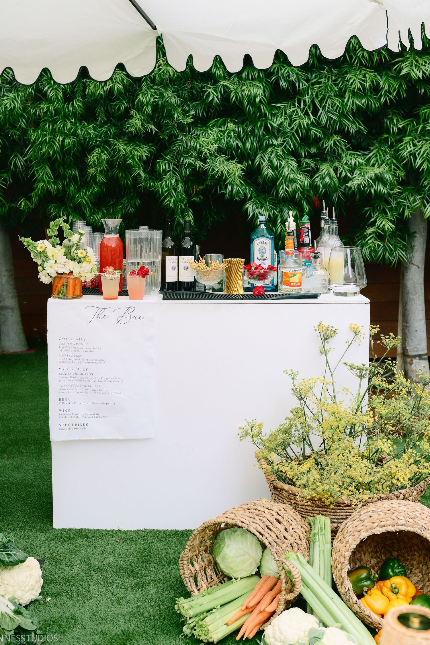 Rustic outdoor bar setup with fresh vegetables and fruits displayed on a white table with garden backdrop.