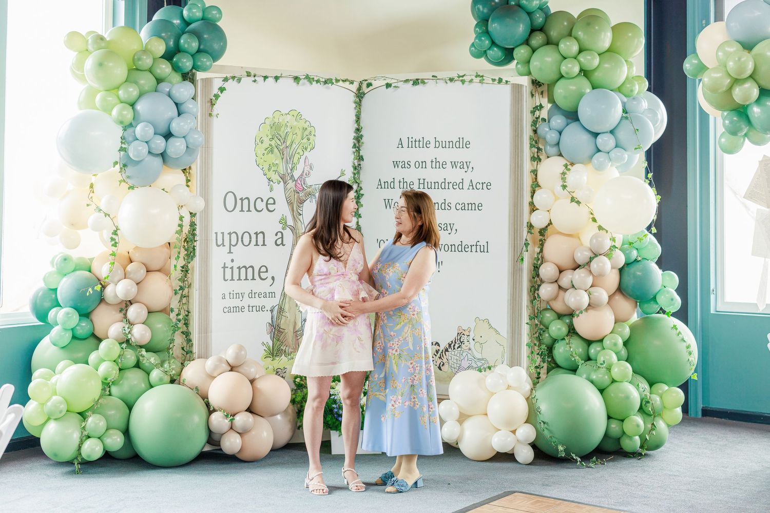 Two people standing in front of a whimsical balloon arch with green and white balloons and storybook backdrop.