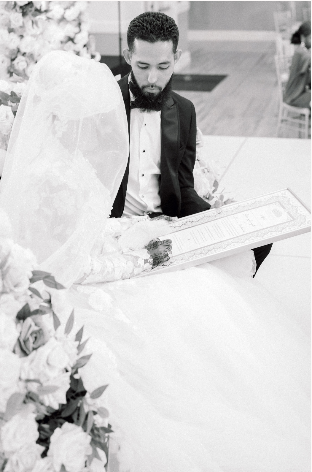 Black and white wedding photo showing couple signing marriage certificate surrounded by floral decorations.
