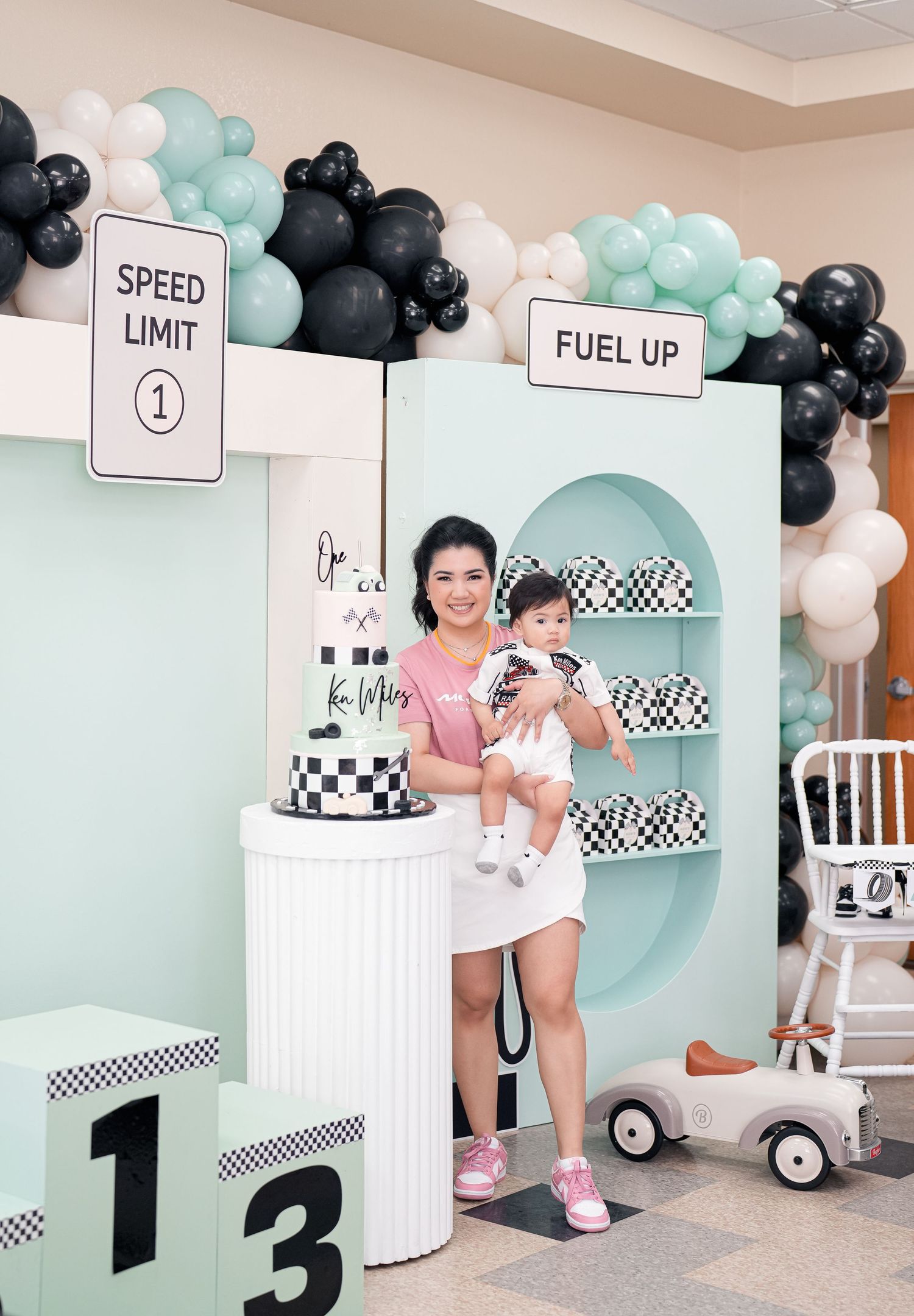 Racing-themed first birthday party setup with mint green walls, checkered decorations, and black and white balloon garlands.