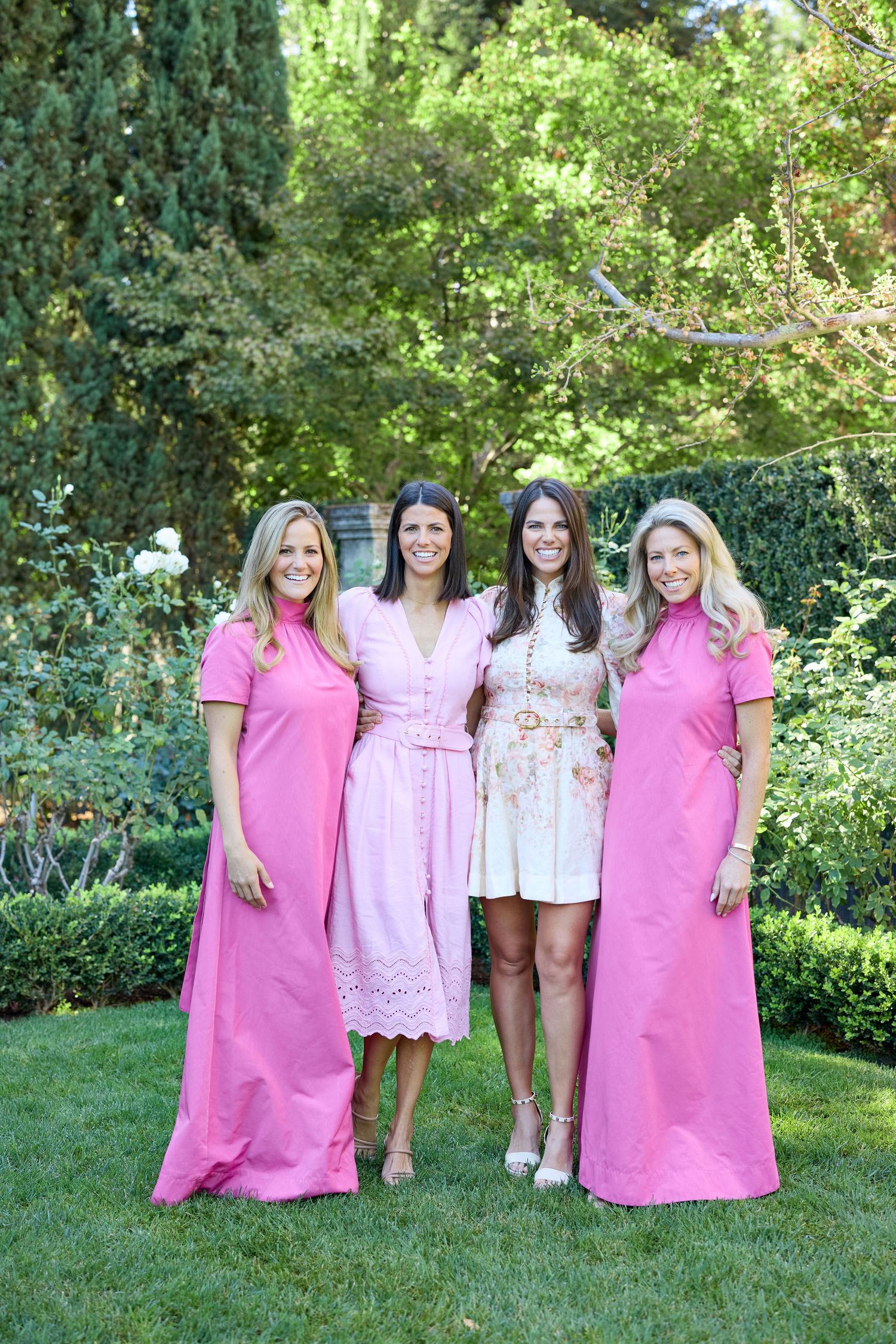 A group wearing coordinated pink dresses poses on a lush green lawn at an outdoor event.