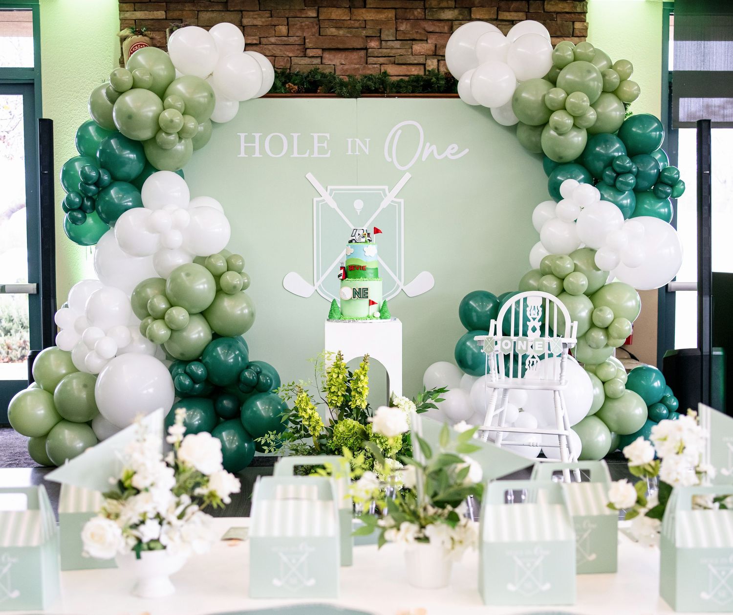 A festive party setup with green and white balloon garlands, decorative chairs, and elegant table settings against a brick wall.