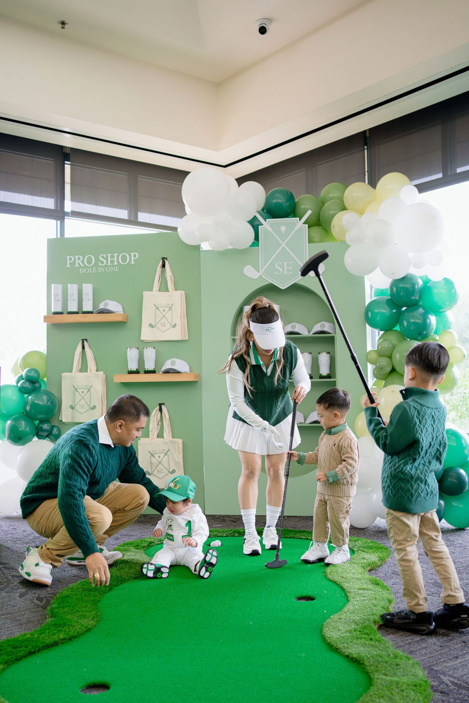 People playing indoor mini golf on a green carpet with white and green balloon decorations overhead.