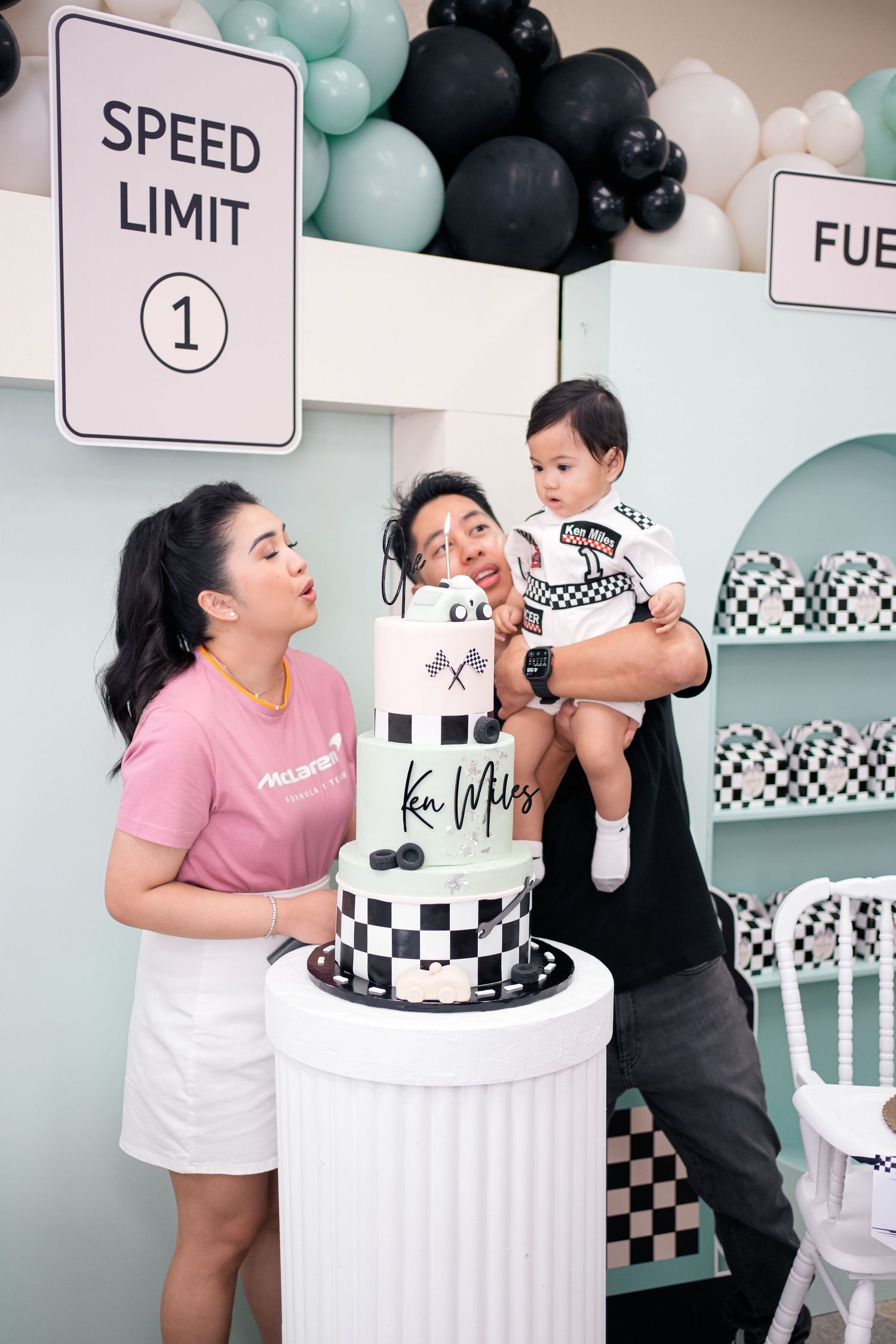 A couple poses playfully with a black and white checkered race car themed birthday cake at a party celebration.