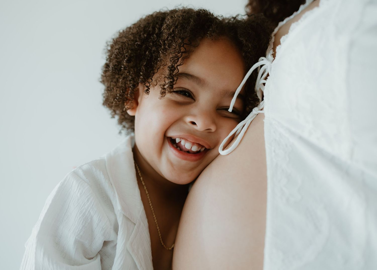 A joyful child with curly hair leans against someone while wearing a white sweatshirt and smiling brightly.