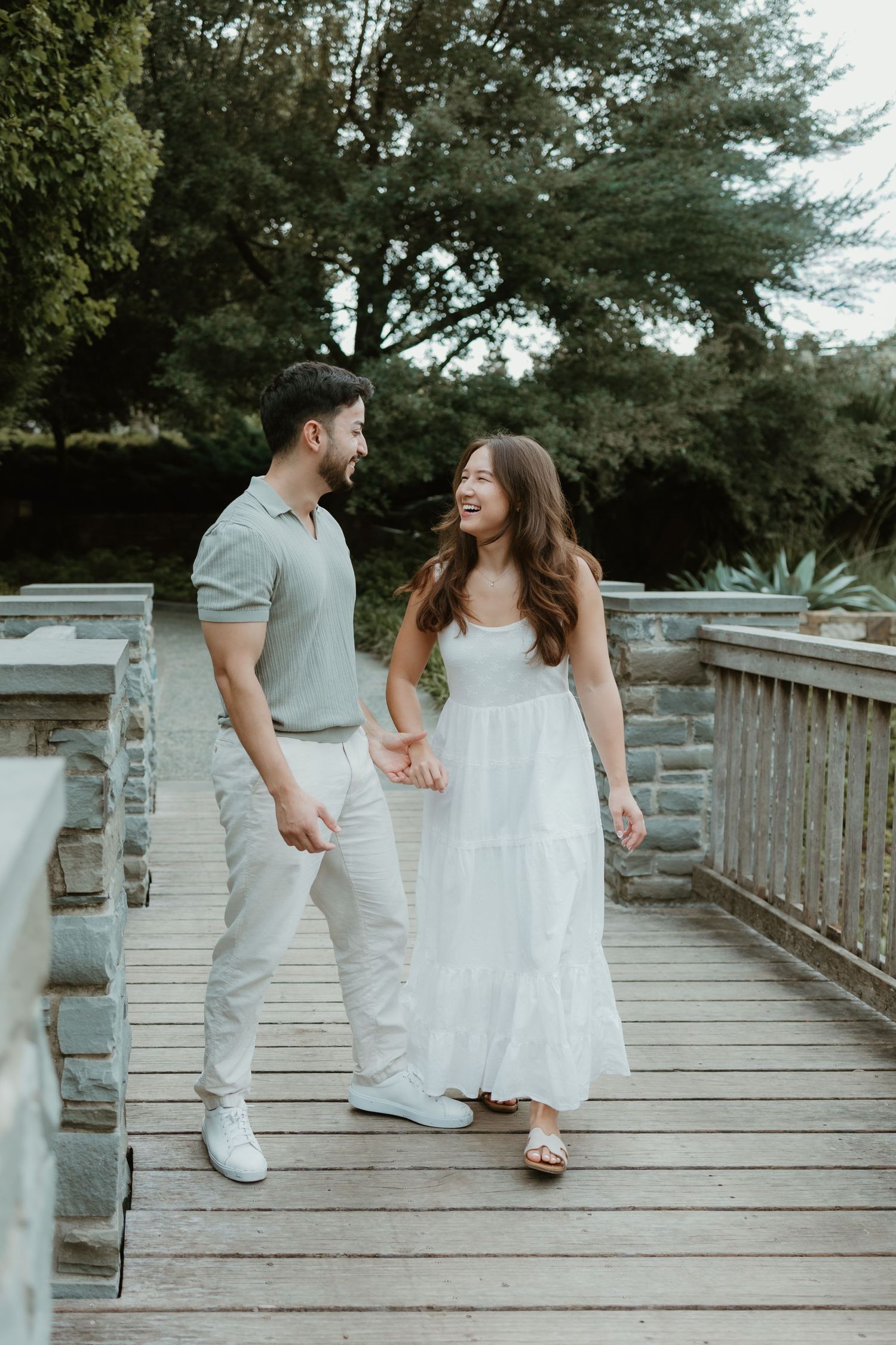 A couple walks hand-in-hand on a wooden bridge, smiling at each other, dressed in white and sage green outfits.