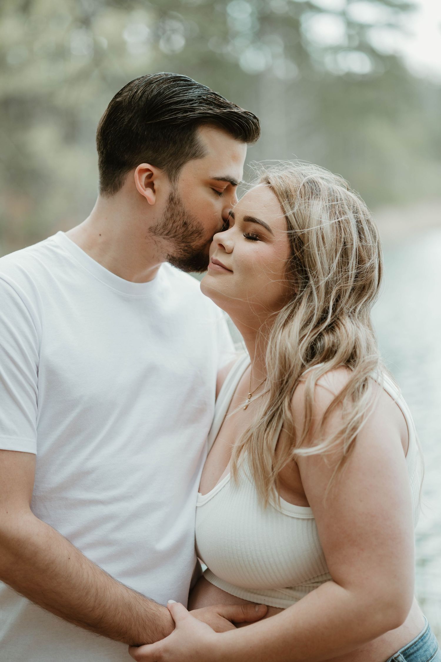 A romantic couple in white outfits share intimate moments during an outdoor photoshoot in a soft, dreamy forest setting.