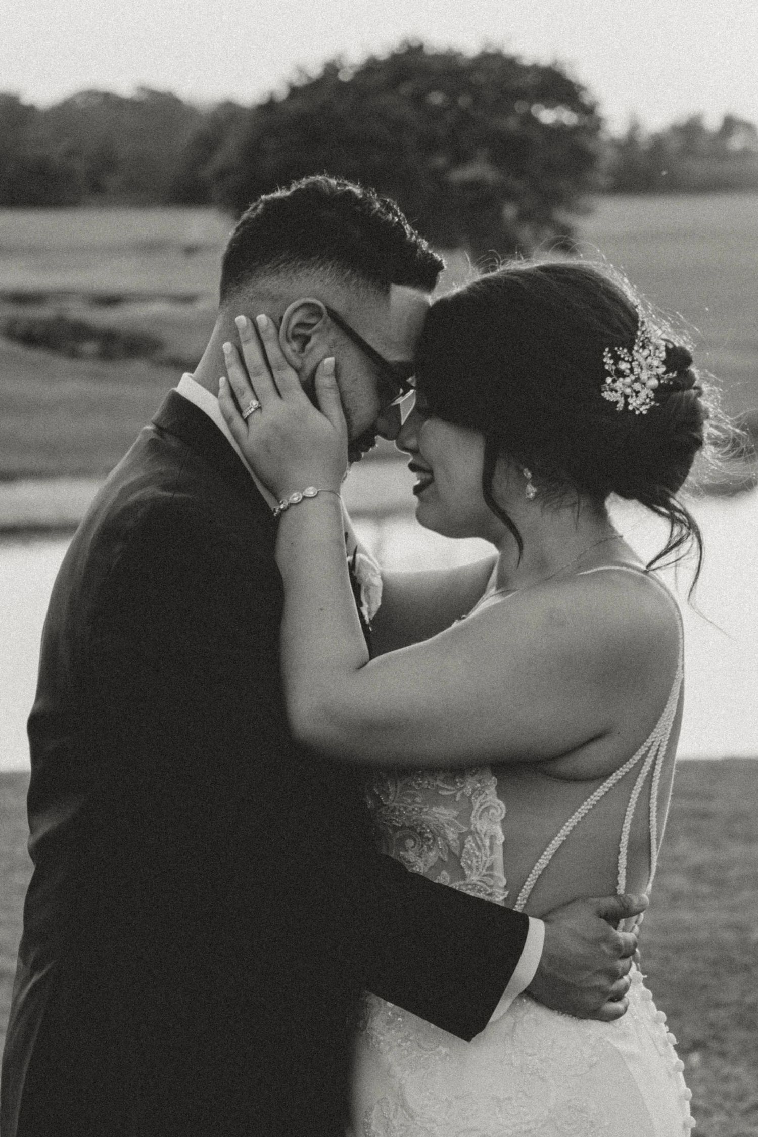 Black and white photo of a bride and groom sharing an intimate moment, nearly kissing, outdoors near a pond.