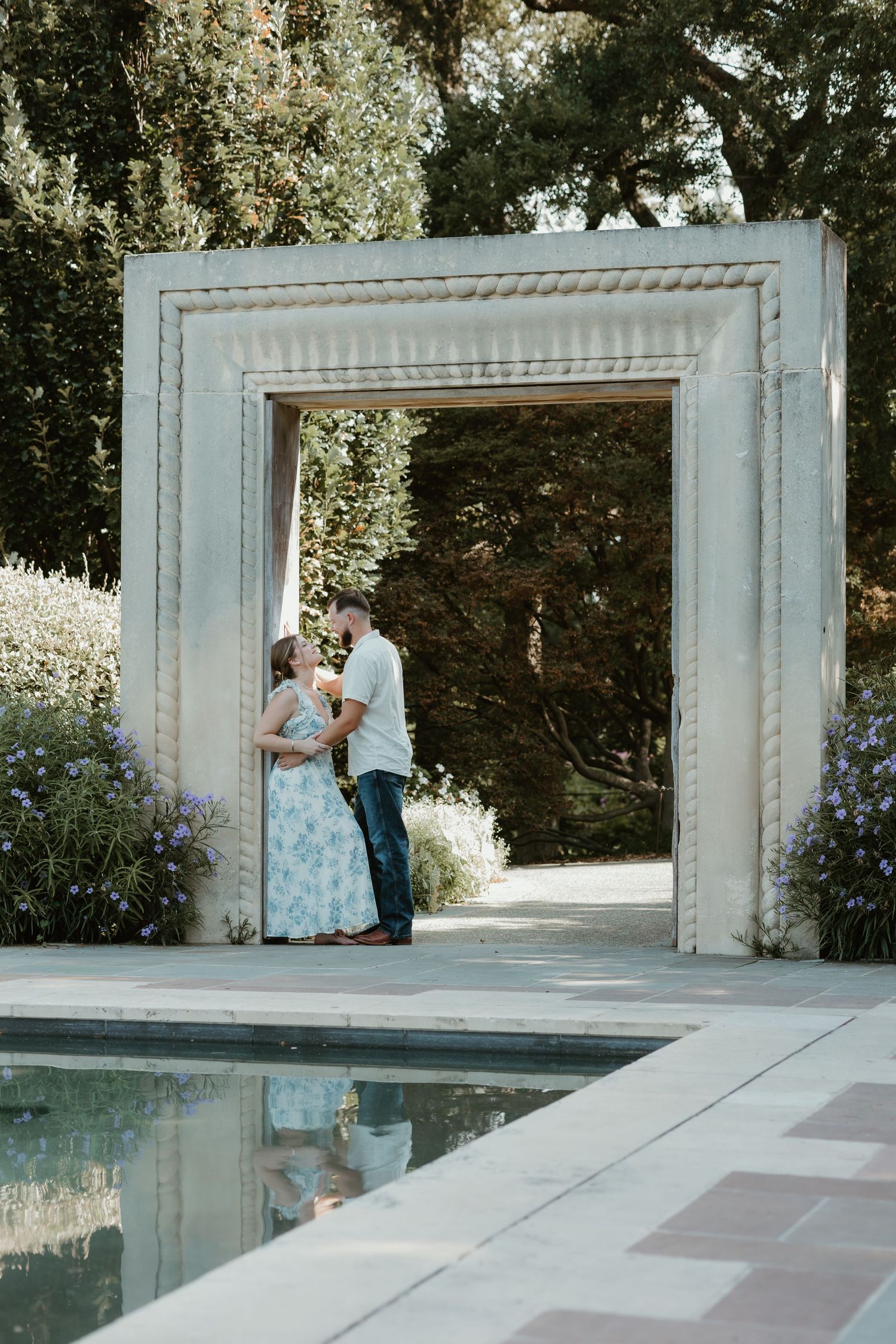 Couple sharing a romantic kiss under a white stone archway beside a reflecting pool in a lush garden.