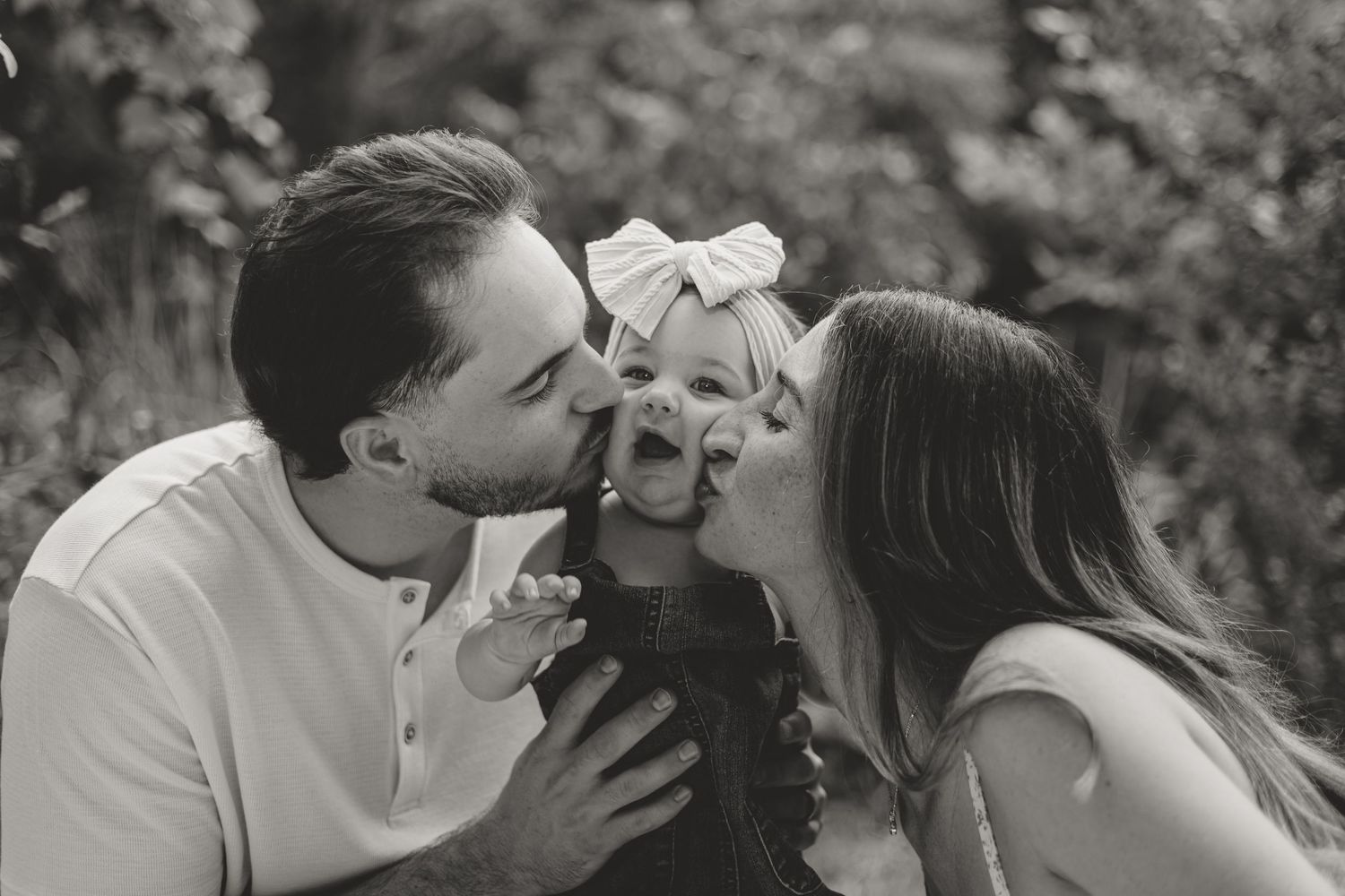 A black and white portrait of parents kissing their baby outdoors in a natural setting.