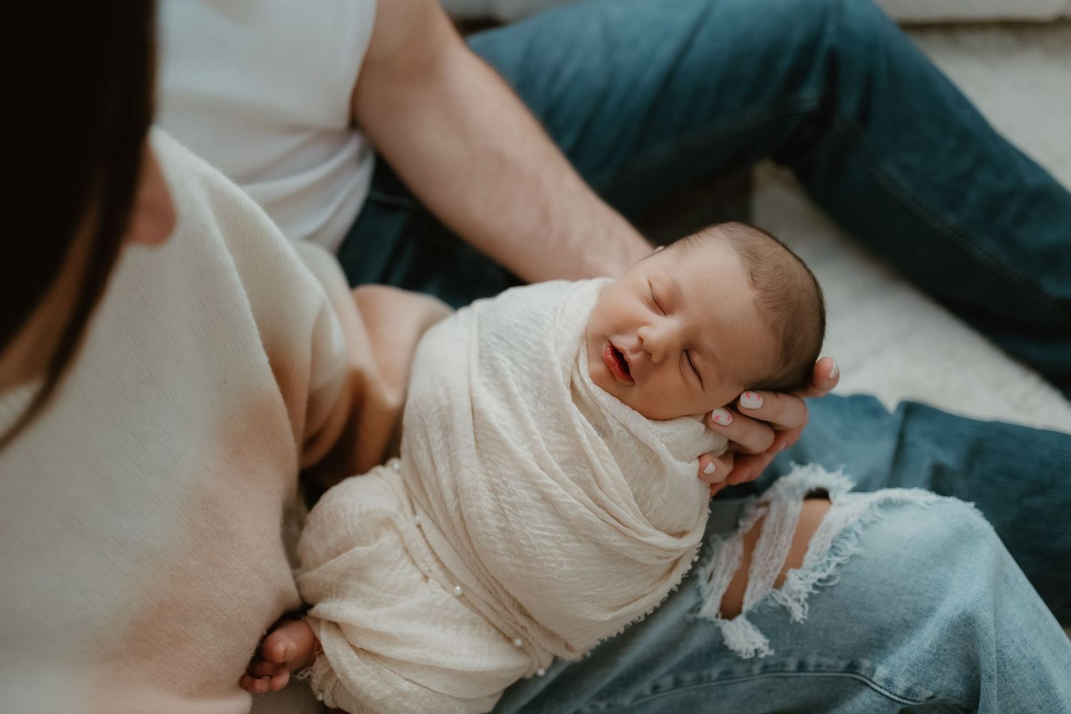 A newborn baby wrapped in white swaddle cloth rests peacefully in their parents lap on a couch.