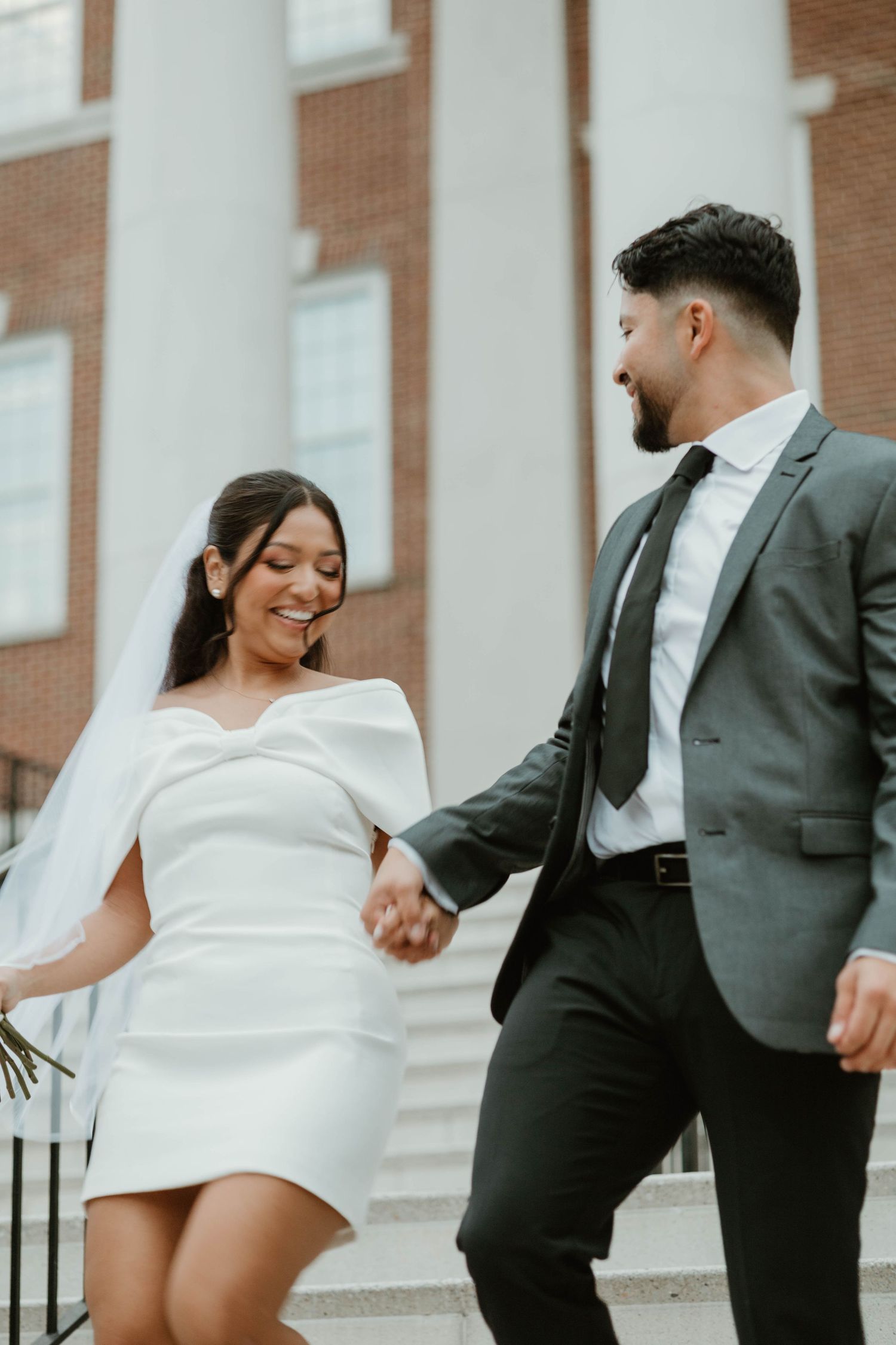 A couple on their wedding day wearing a short white dress and gray suit descending steps together hand in hand.
