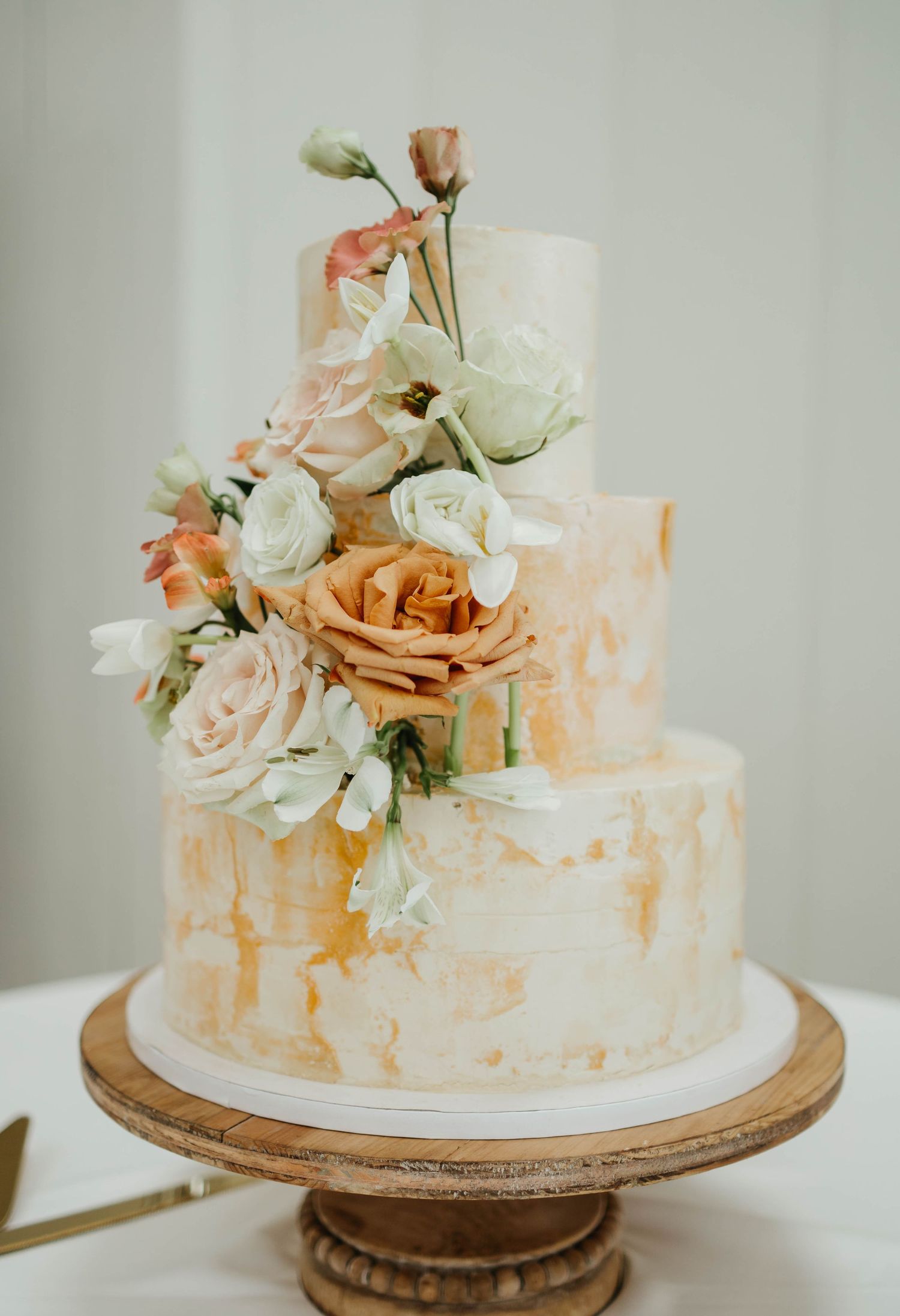 Elegant three-tier wedding cake with gold marble finish adorned with fresh peach and white roses on wooden cake stand.