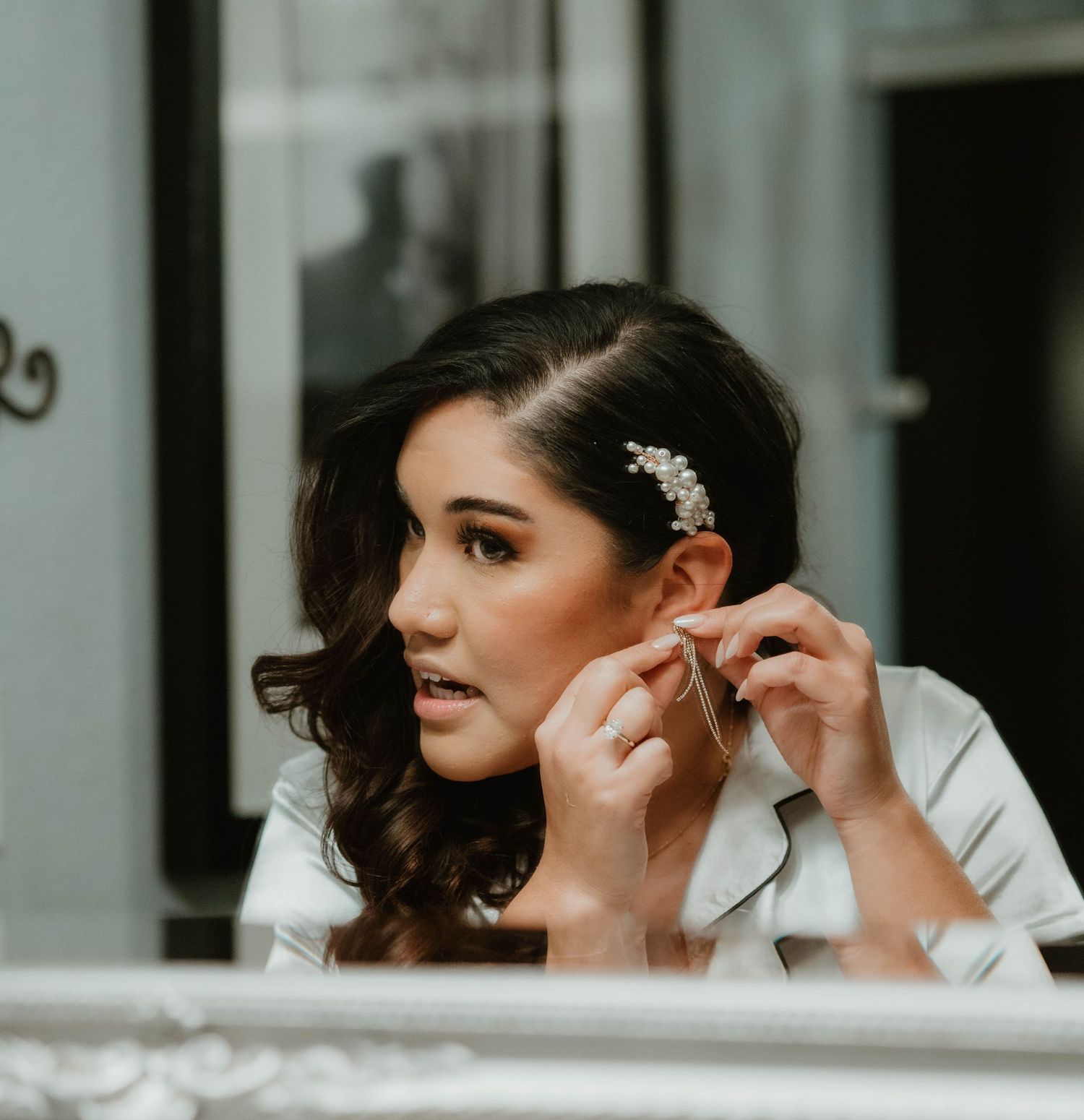 Someone in a white top puts on earrings while getting ready, with styled dark wavy hair and delicate hair accessories.