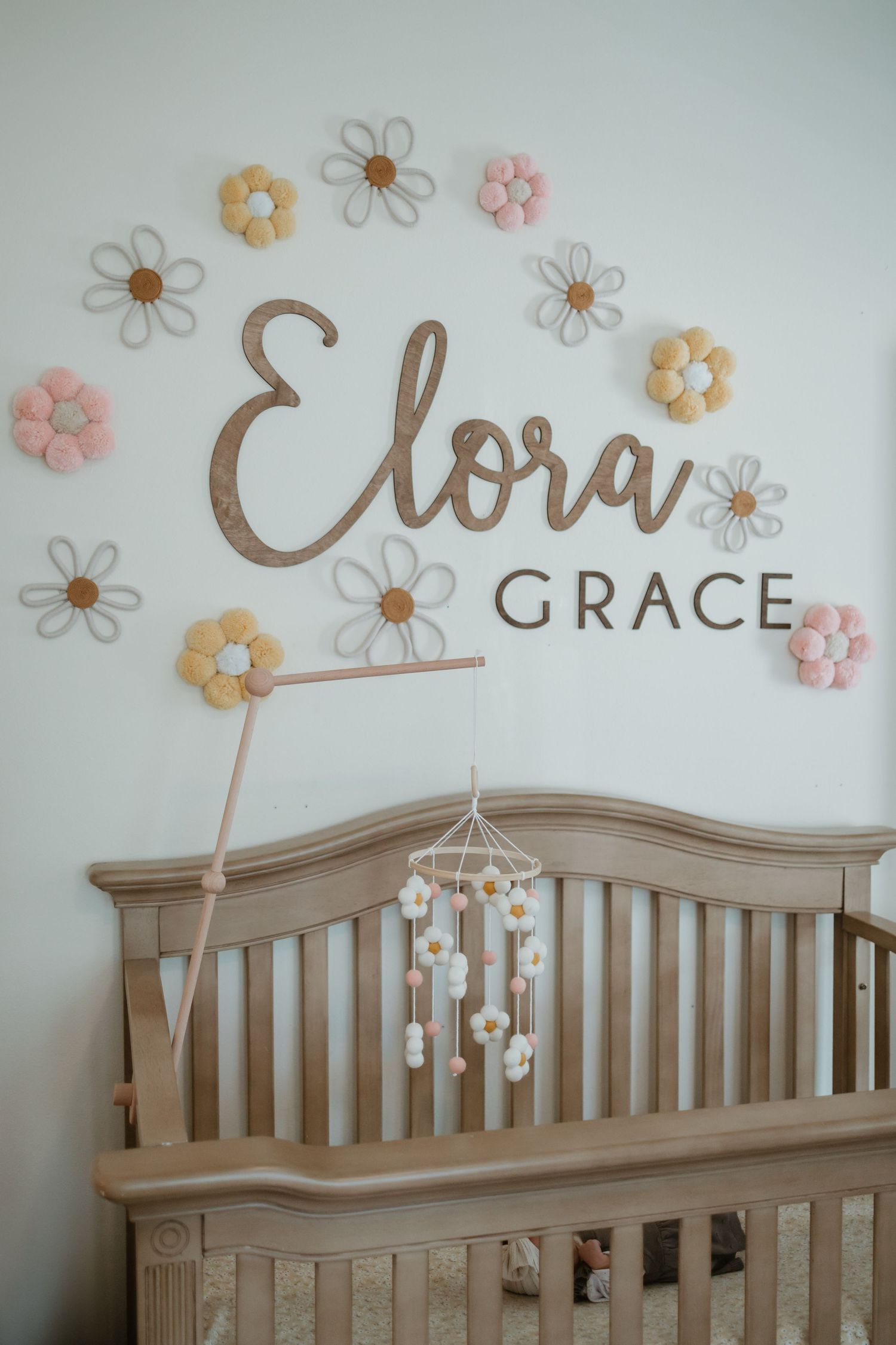 Baby nursery crib with Elora Grace wooden name sign and daisy flower wall decor in pink, white, and yellow.
