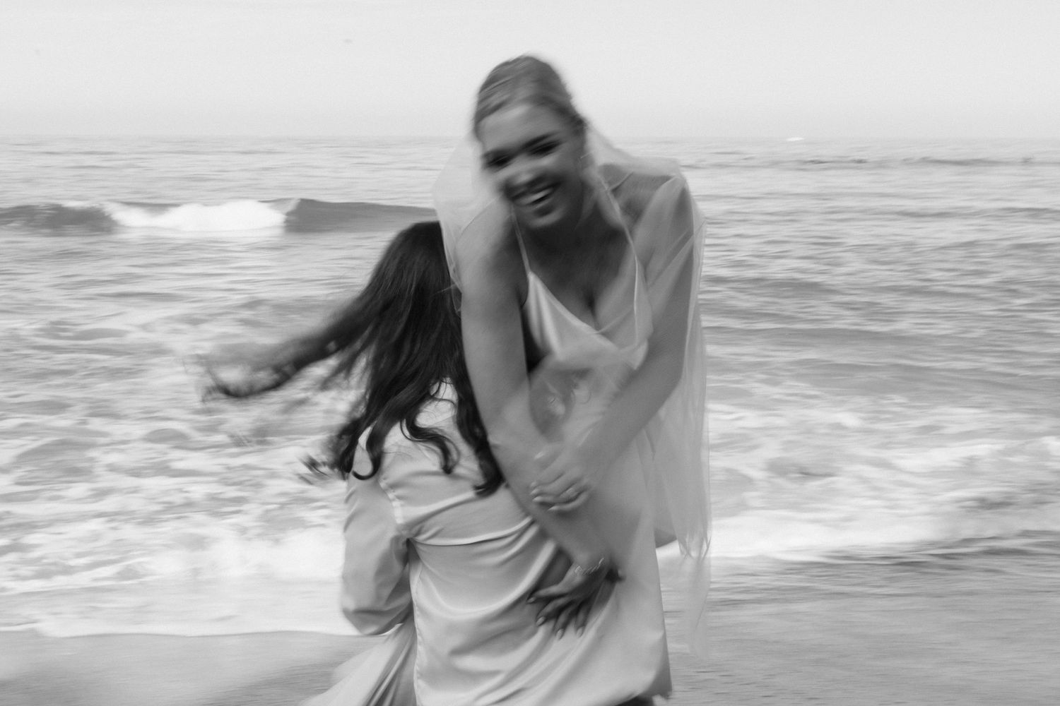 A couple in casual white clothing plays and laughs together on a misty beach shoreline during golden hour.