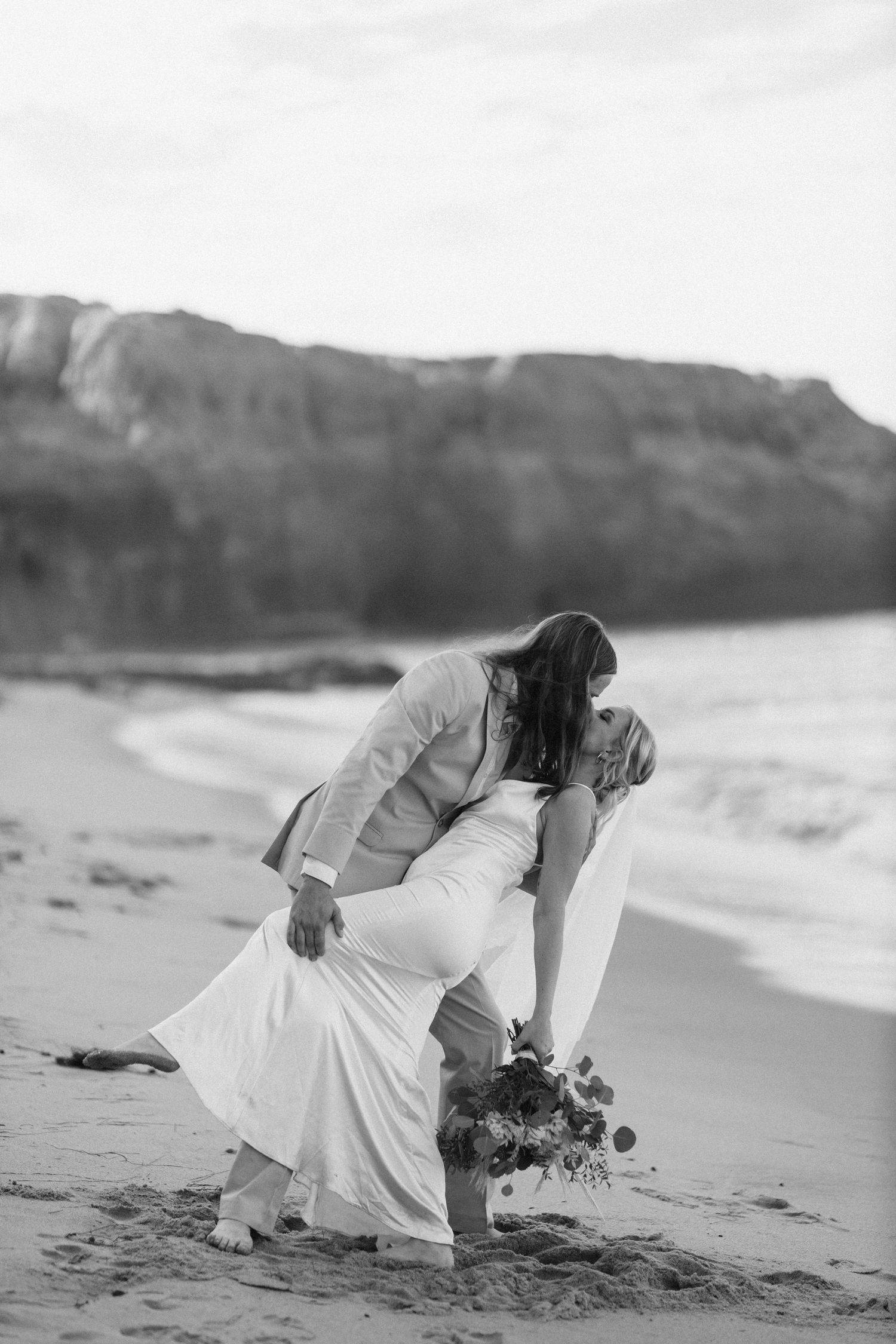 Bride in flowing white wedding dress bends down to pick up her bouquet on a scenic beach at sunset with cliffs behind.