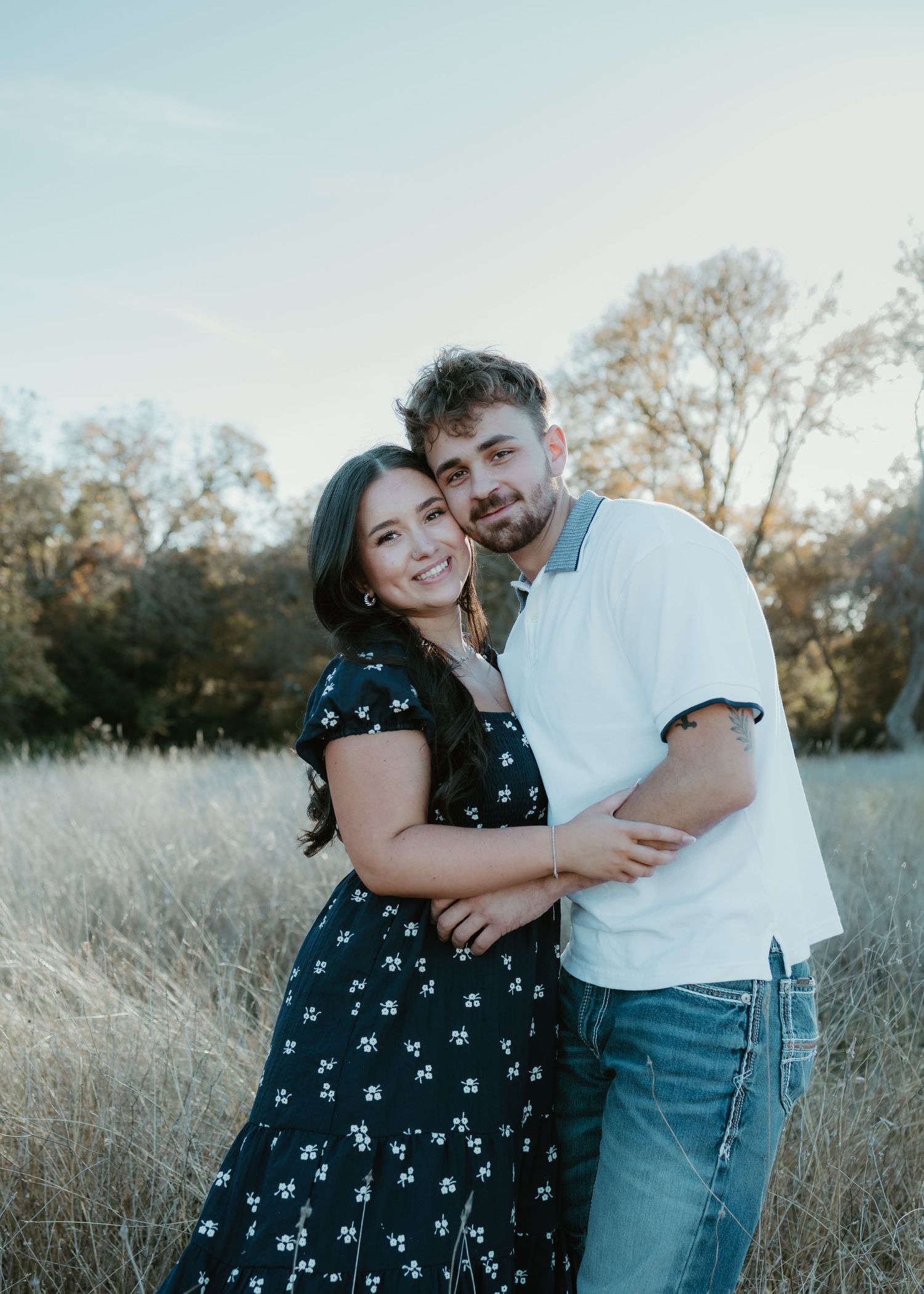 Couple embracing in a golden field, woman in floral dress and man in white shirt and jeans, smiling outdoors.