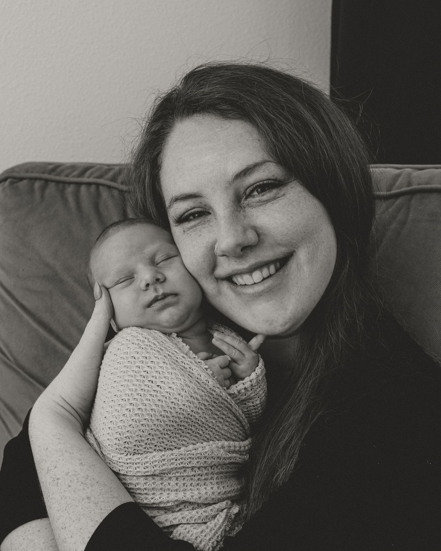 Black and white photo of a smiling woman holding a sleeping newborn baby wrapped in a knit blanket.