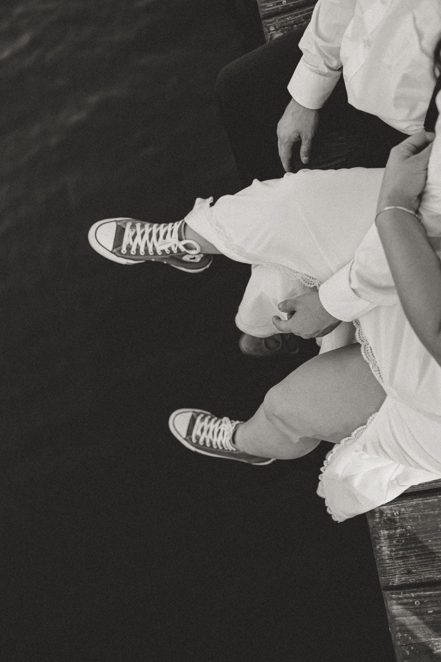 Black and white photo of two people sitting on a dock wearing Converse sneakers and white pants near water.