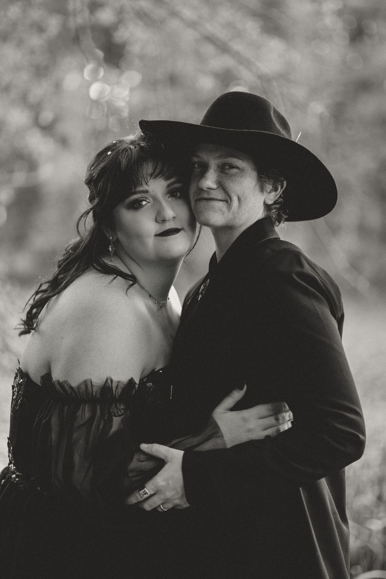 Black and white photo of a couple embracing, woman in off-shoulder dress, man in black suit and wide-brim hat.