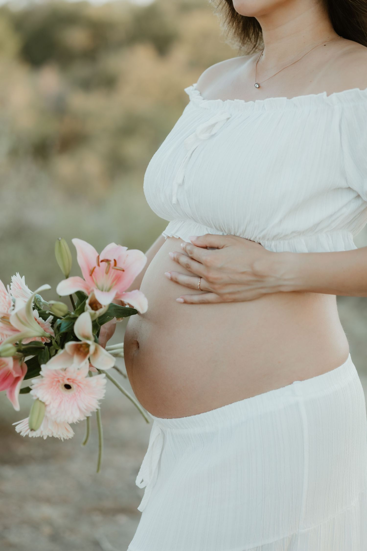 A pregnant woman in a white dress holds pink and white flowers against her baby bump in a maternity photo series.