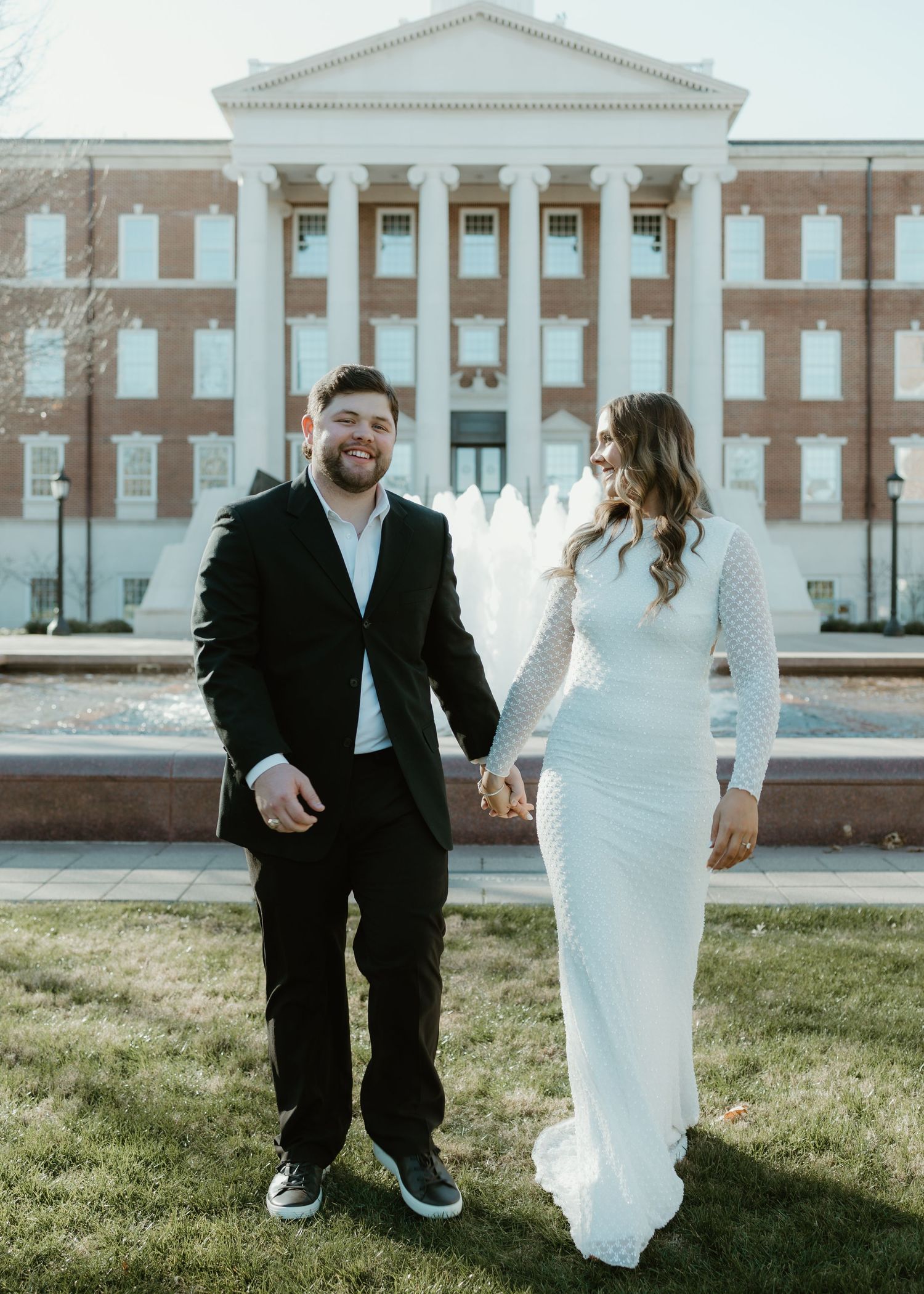 Couple holding hands smiling in front of brick building with fountain, man in black suit, woman in white fitted gown.