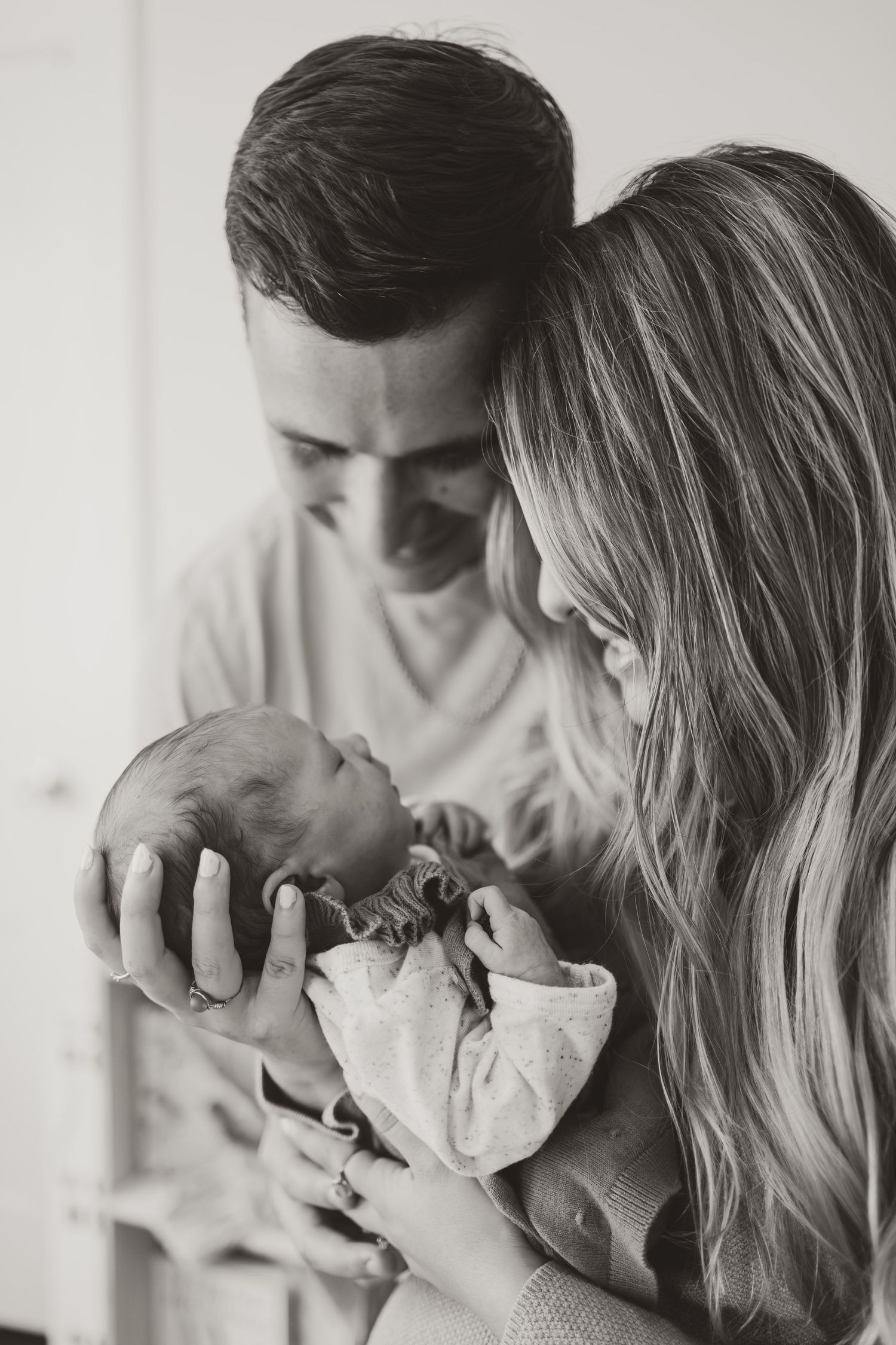 Black and white photo of new parents lovingly gazing at their newborn baby held gently in their hands.