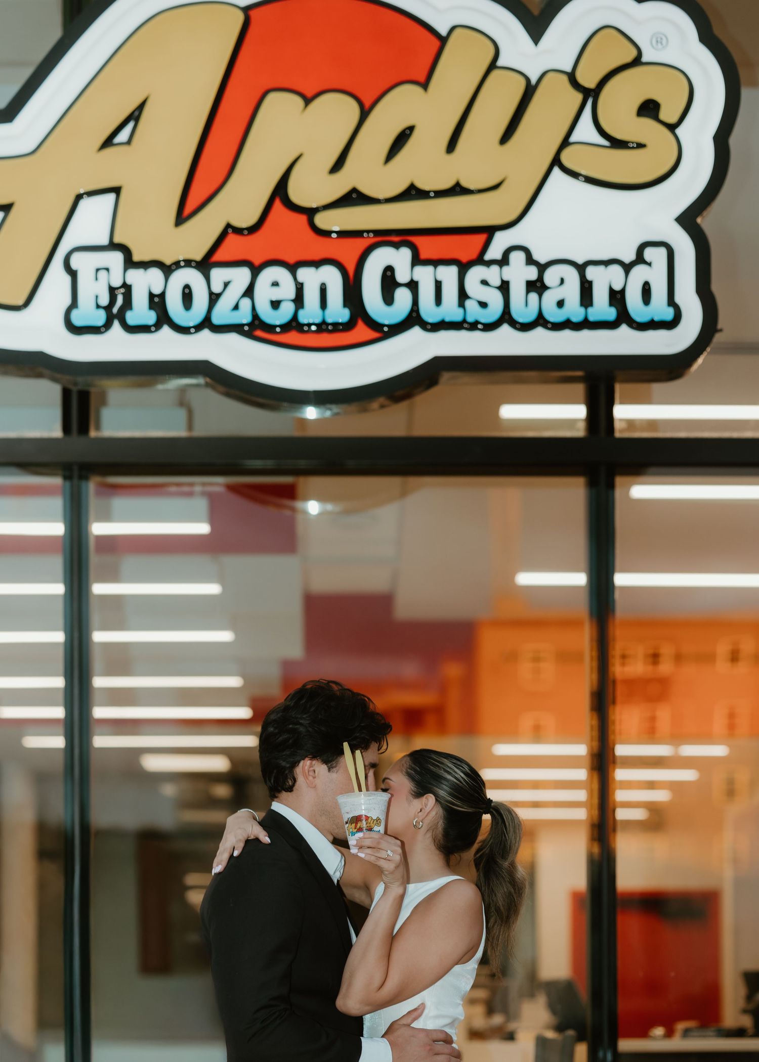Couple in wedding attire sharing a kiss holding frozen custard outside Andy's Frozen Custard shop.