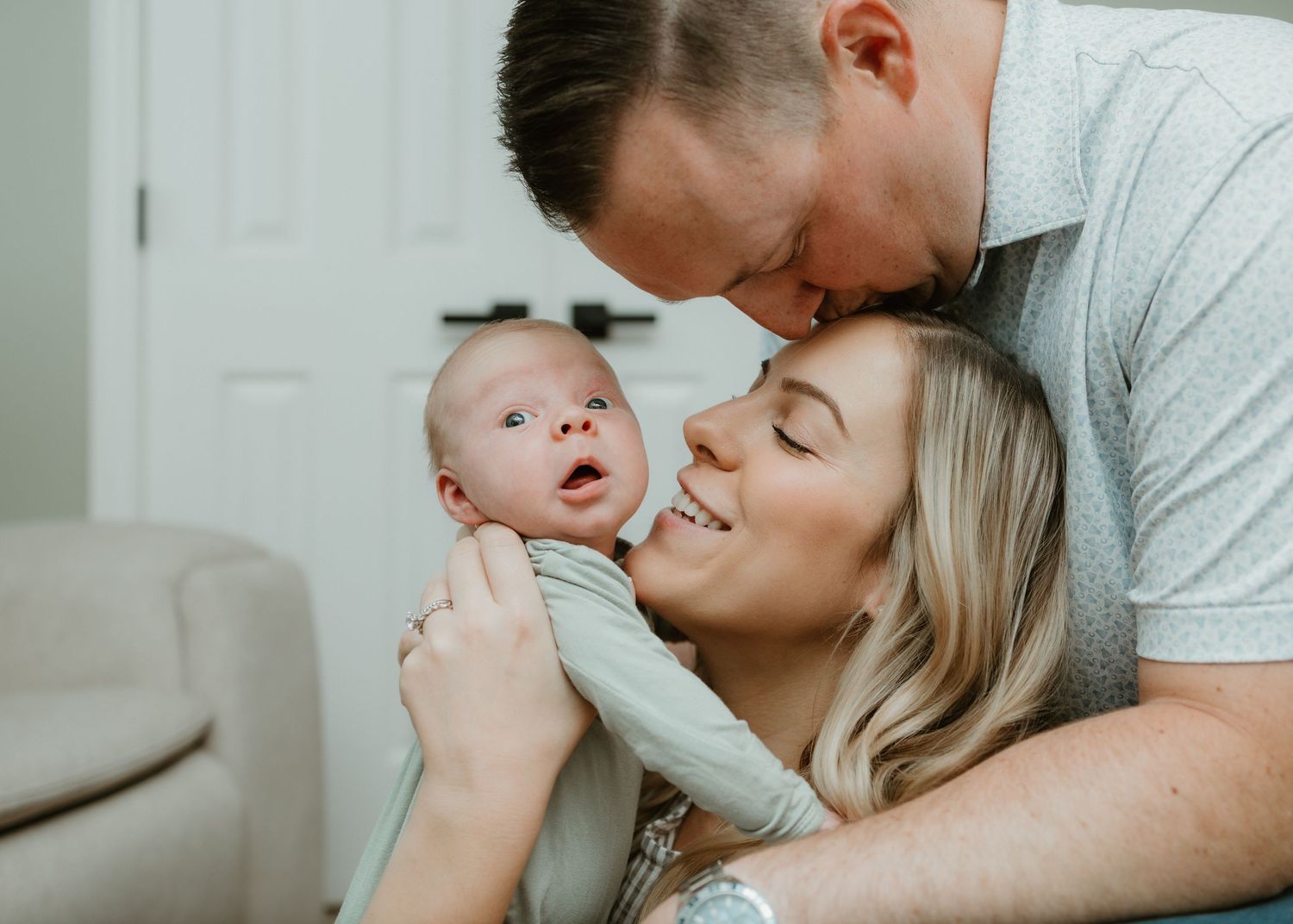 A loving couple shares a tender moment kissing their newborn baby in a bright, cozy home setting.