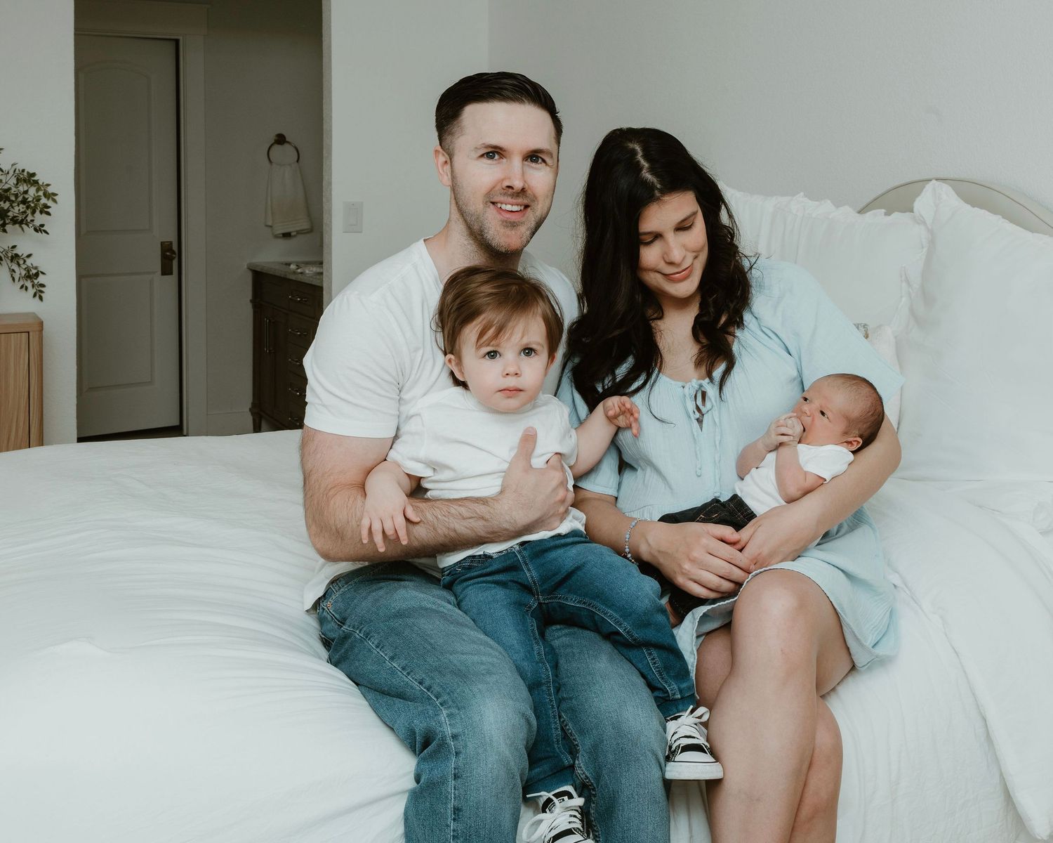 Happy family of four sitting on a white bed, parents holding two young children including a newborn baby.