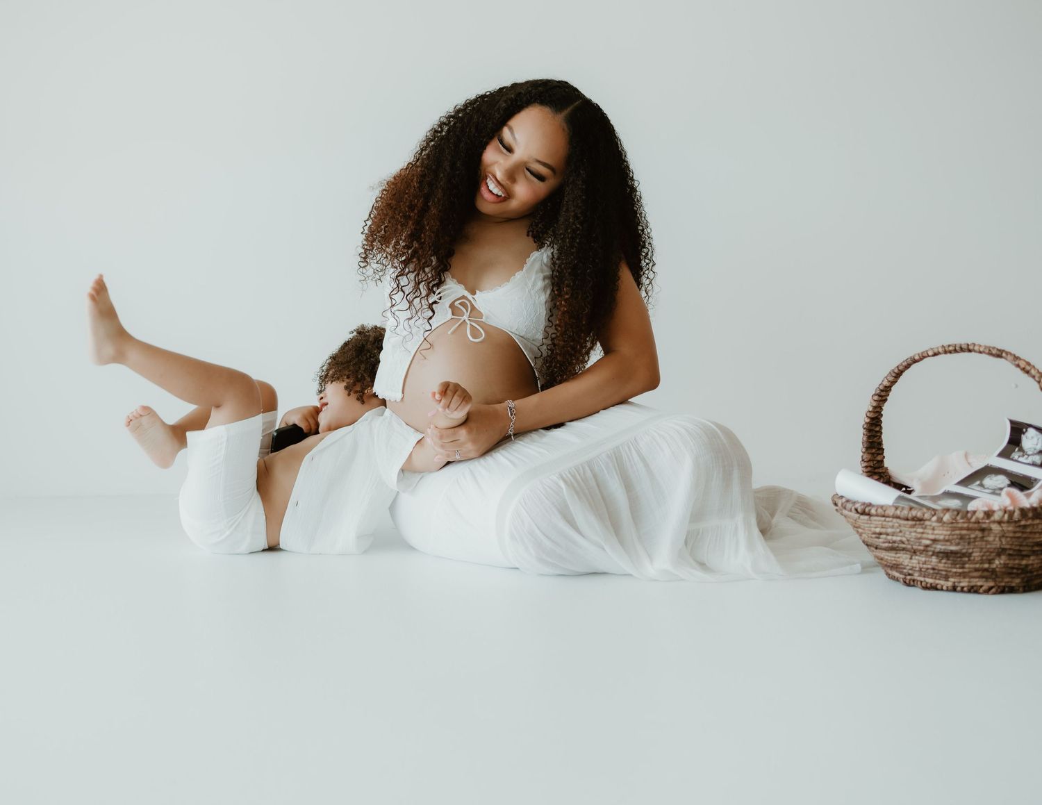 Person in white outfit sitting on light background with natural curly hair and wicker basket beside them.