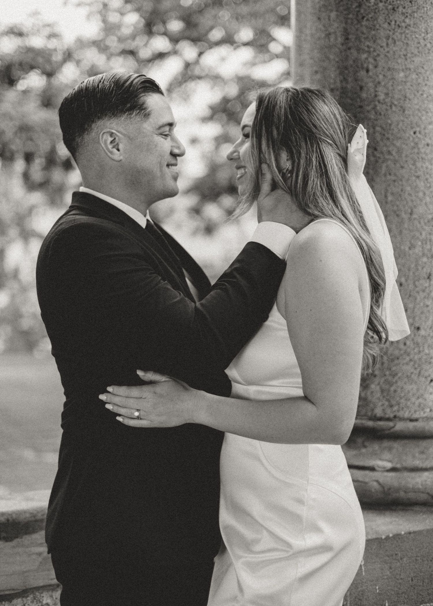 Black and white romantic wedding photo of a couple embracing outdoors by a stone pillar.