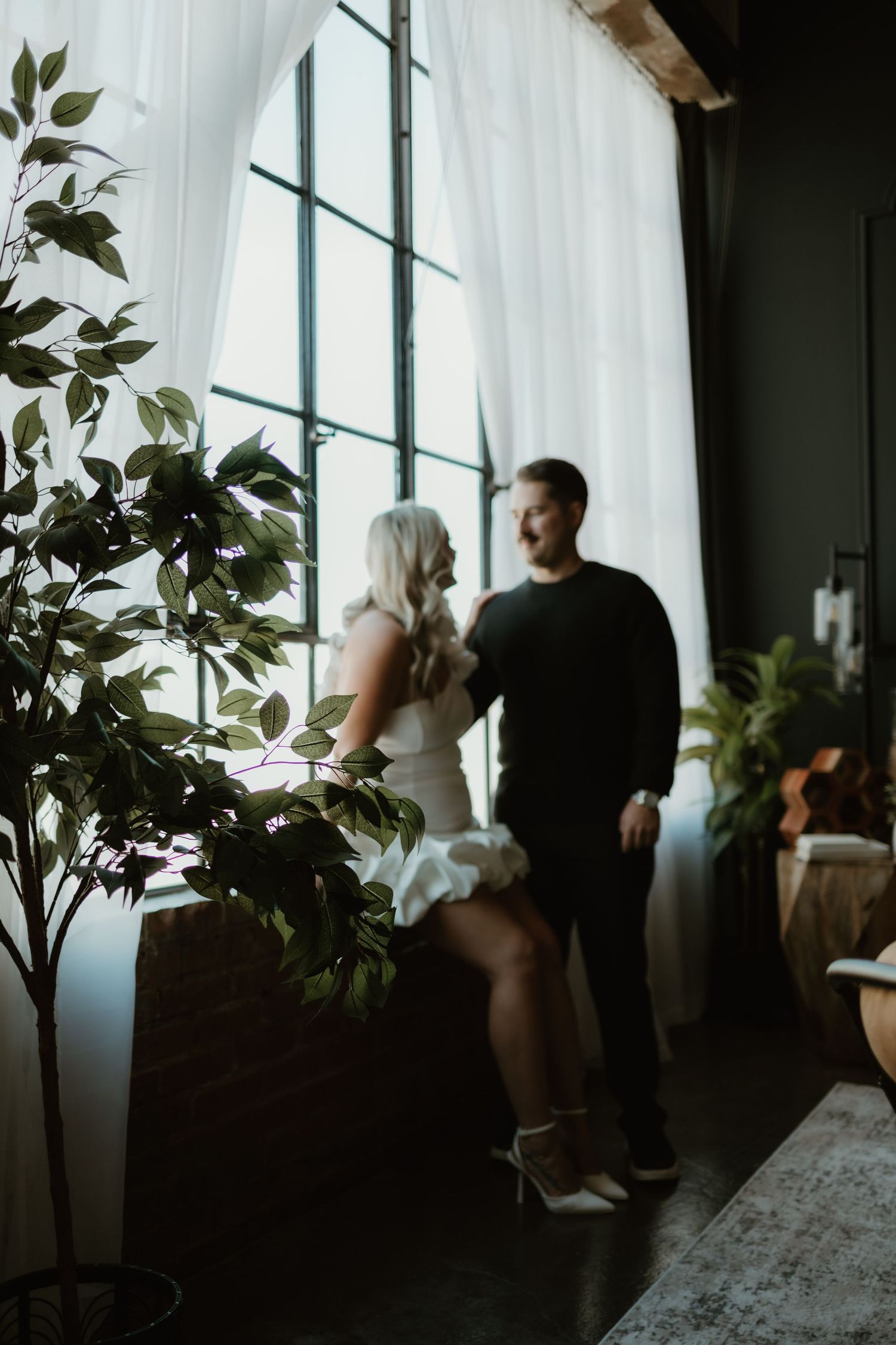 A couple gazes out large windows in moody lighting, standing beside indoor plants in an intimate indoor setting.