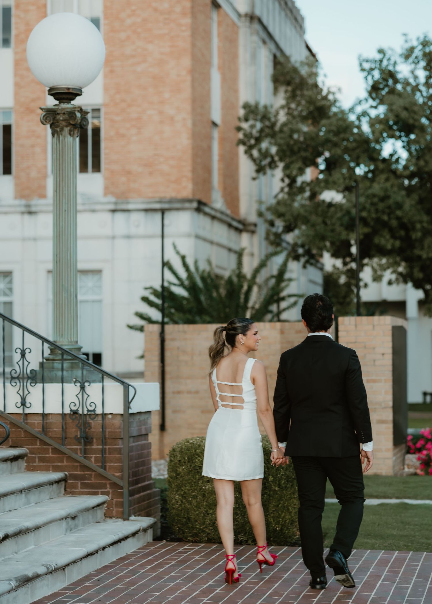 Couple in wedding attire holding hands walking toward brick building, woman in white mini dress with red heels, man in black suit.