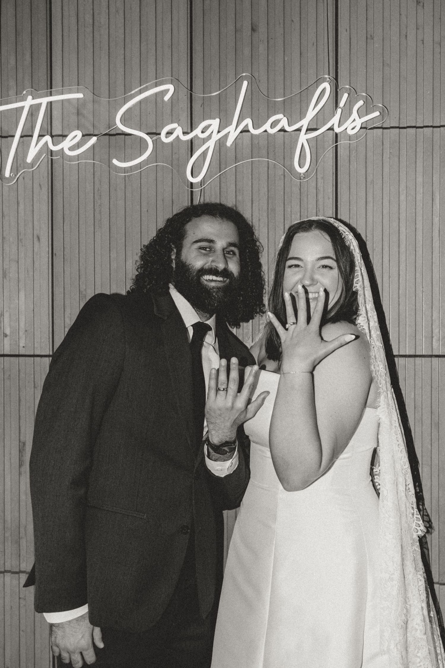 Newlyweds The Saghafis pose joyfully in black and white wedding photo, bride in veil showing rings, groom in suit.
