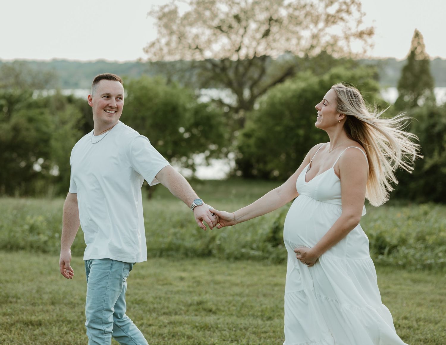 Smiling couple holding hands in a green field, pregnant woman in white dress with wind-blown hair during maternity photoshoot.