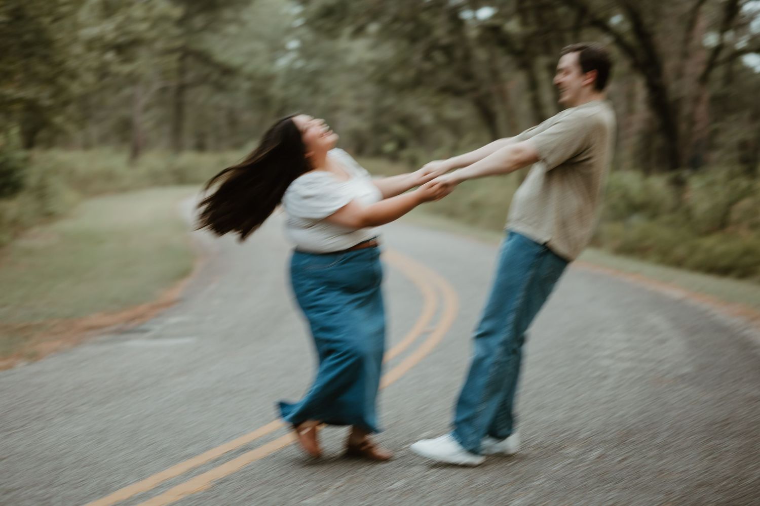 Two people playfully spin and dance together on an empty tree-lined road in a whimsical outdoor moment.
