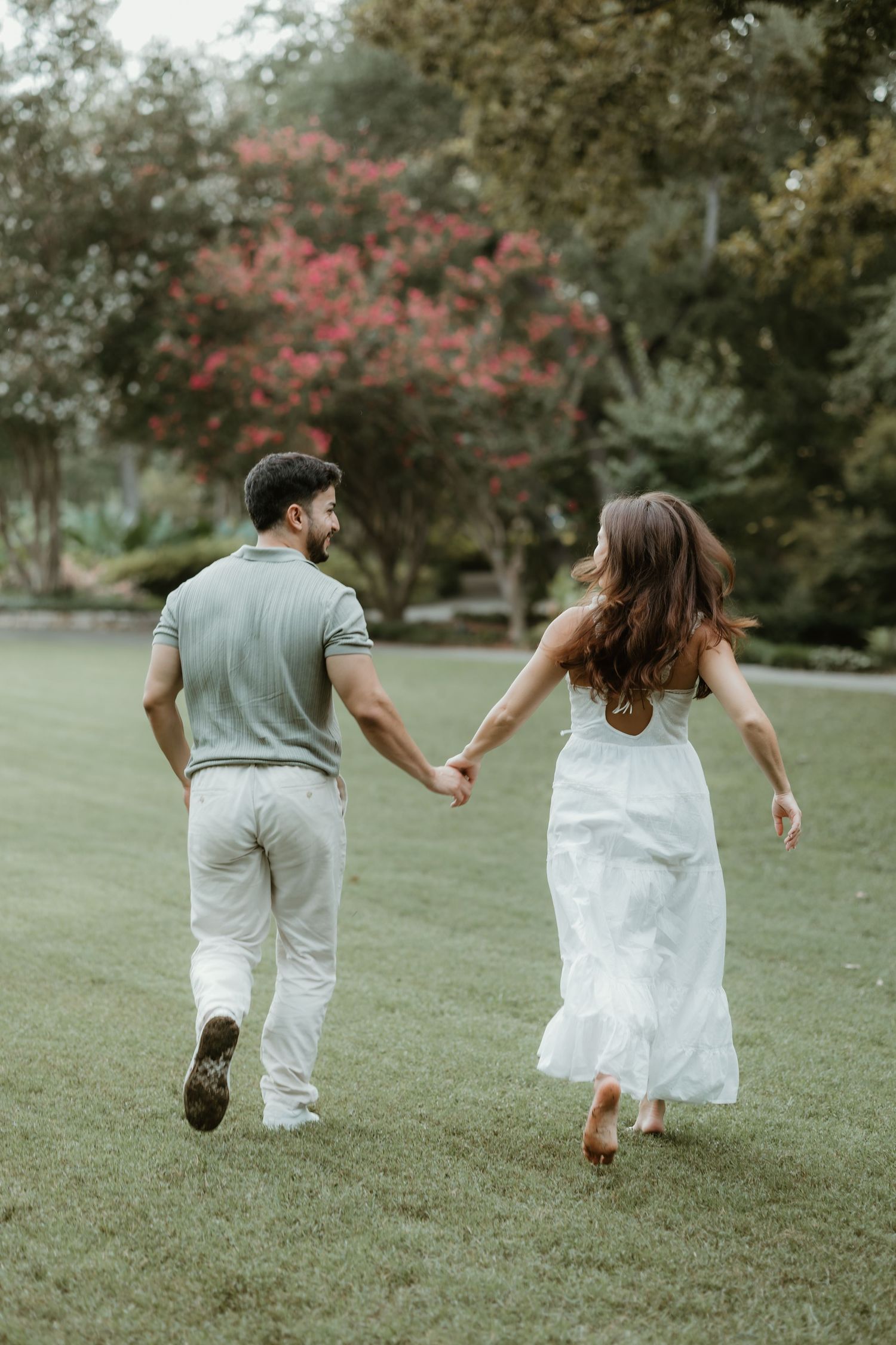 A couple in matching white and gray outfits runs playfully through a lush green park on a sunny day.