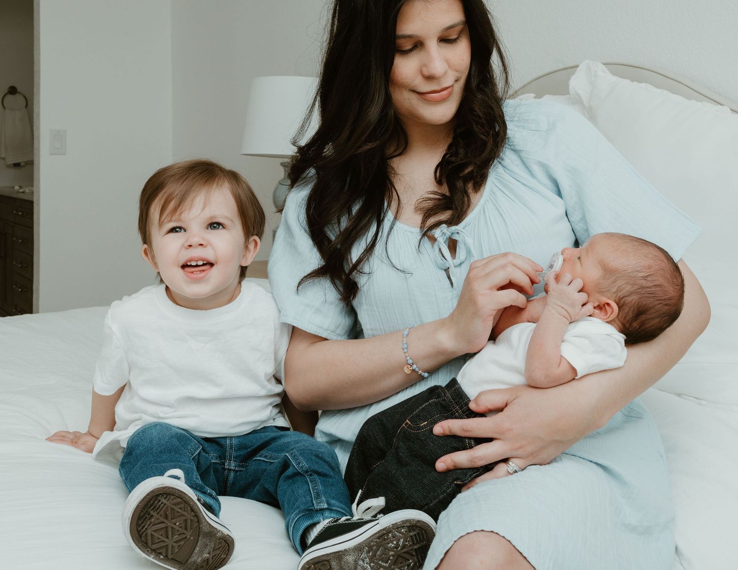 Mother in blue dress holds newborn baby while toddler sits smiling beside her on white bed.