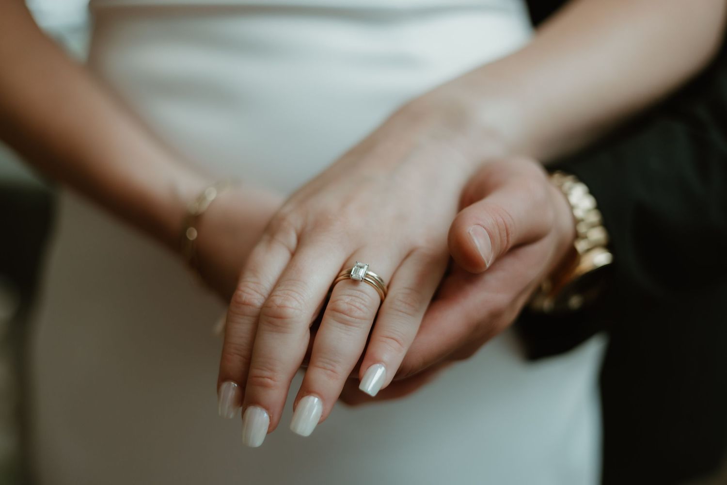 Close-up view of hands wearing an engagement ring with neutral manicured nails and gold bracelet.