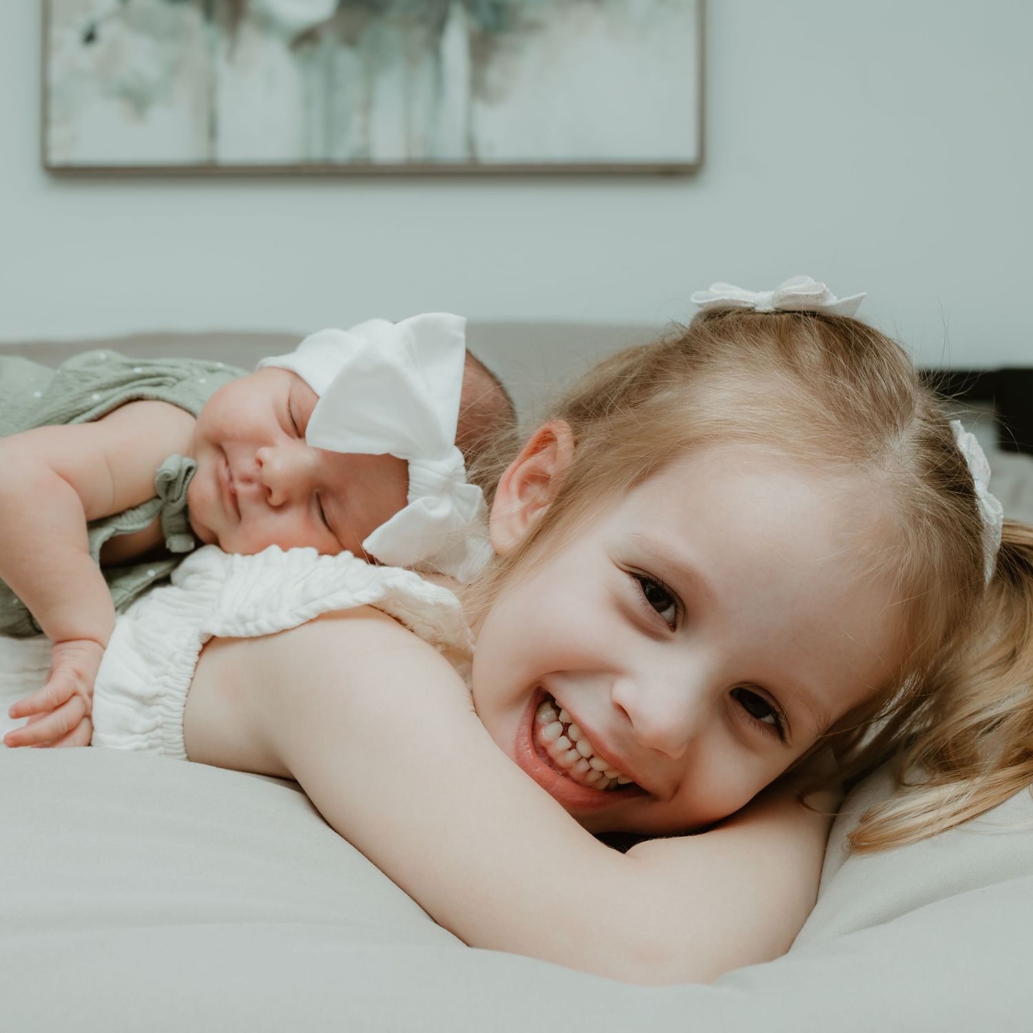 Smiling toddler girl lying on a bed next to a sleeping newborn baby wearing a white bow headband.