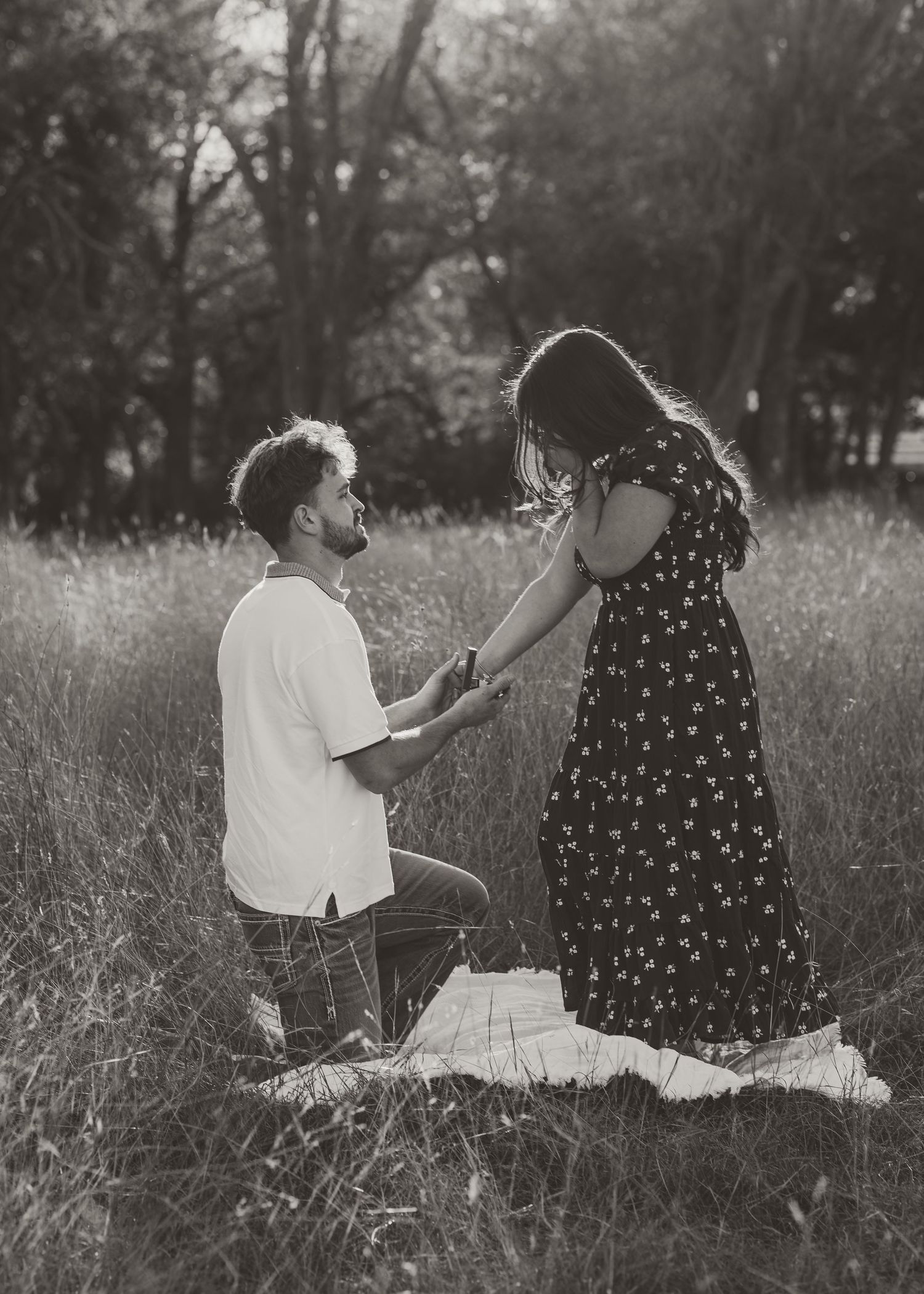 Man kneeling on one knee proposing to woman in floral dress in a grassy field, black and white photo.