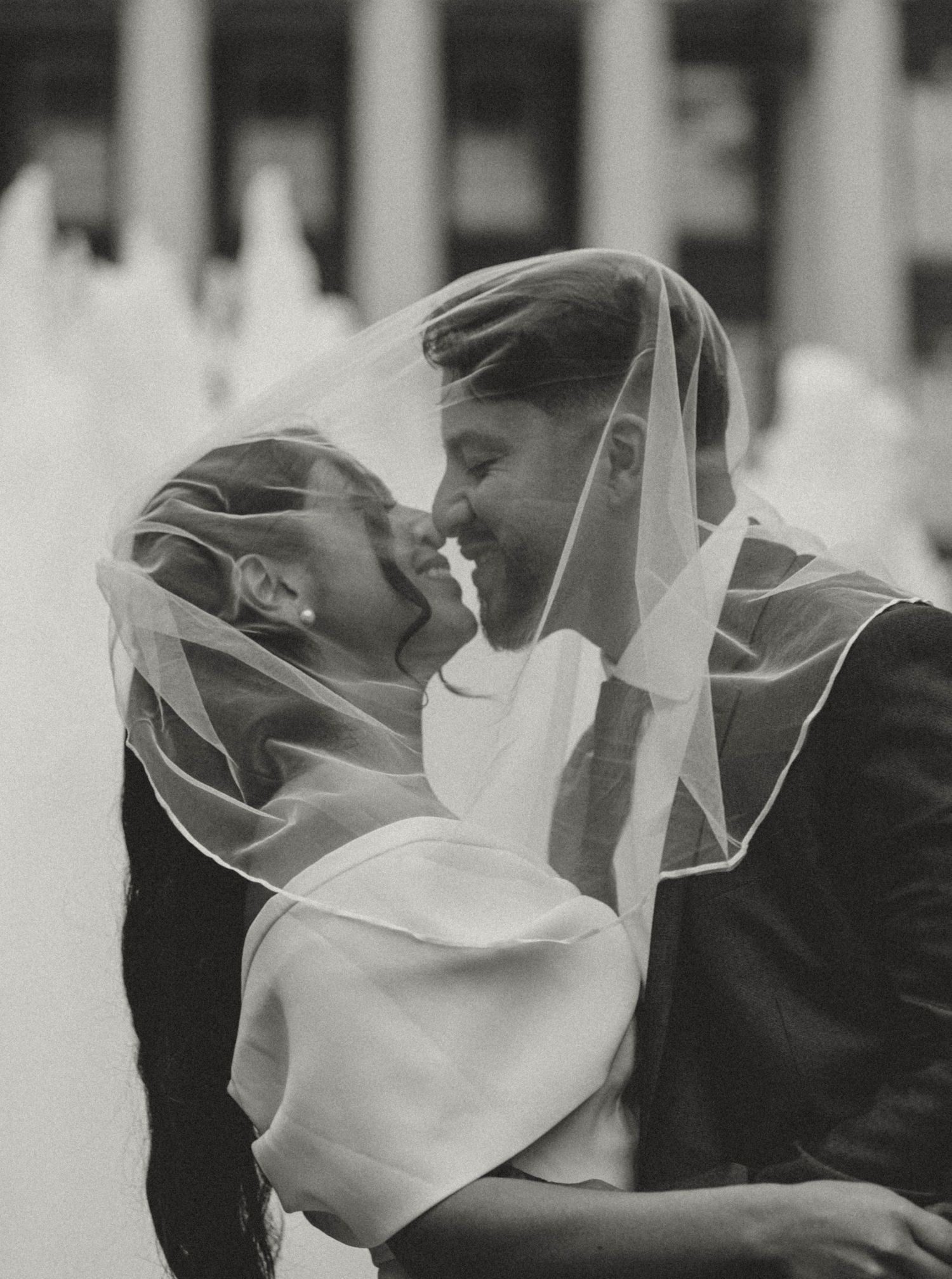 Black and white photo of a bride and groom sharing a romantic kiss under a wedding veil near a fountain.