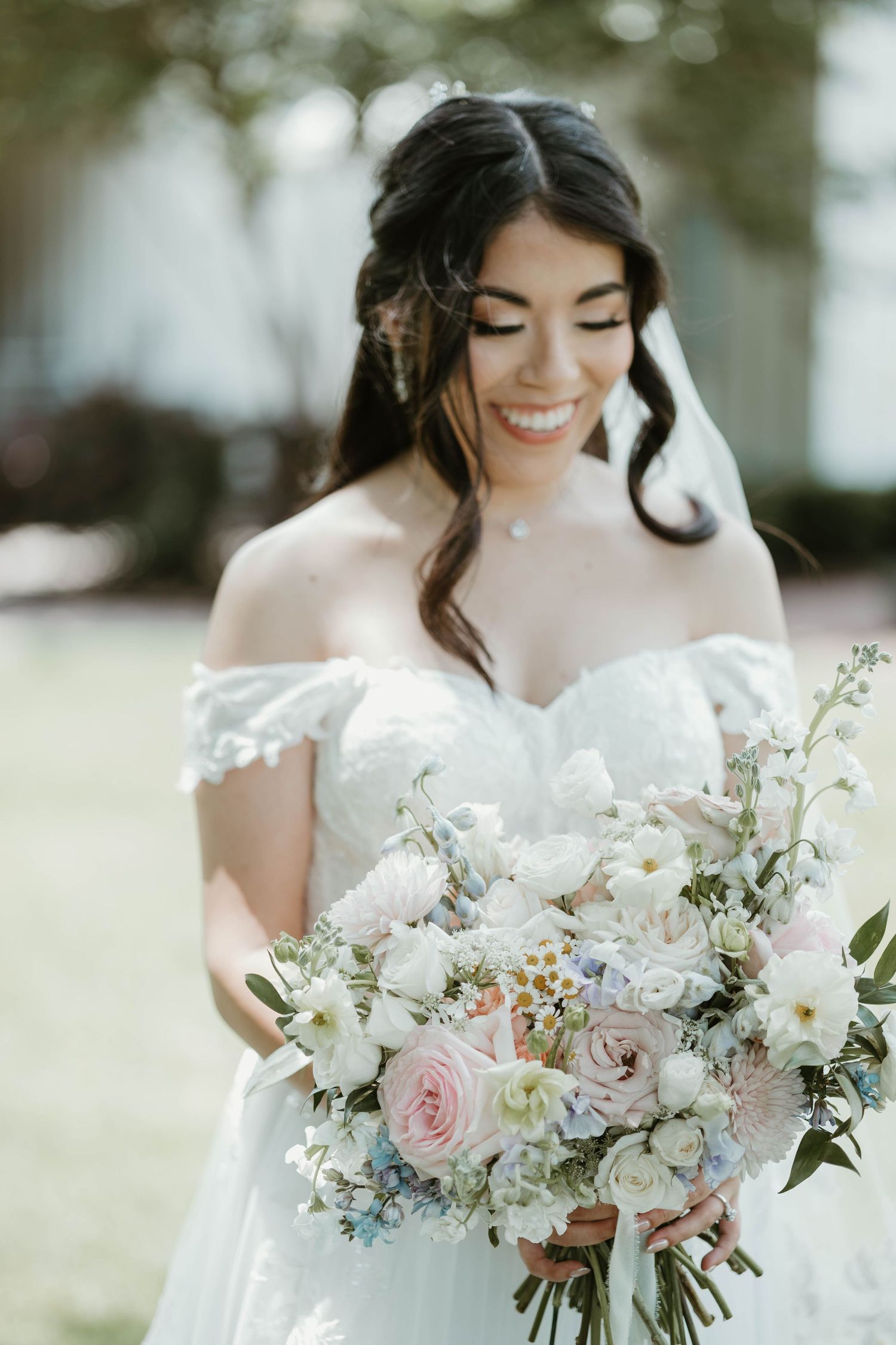 Smiling bride in off-shoulder lace gown holding a lush bouquet of white and blush pink flowers on her wedding day.