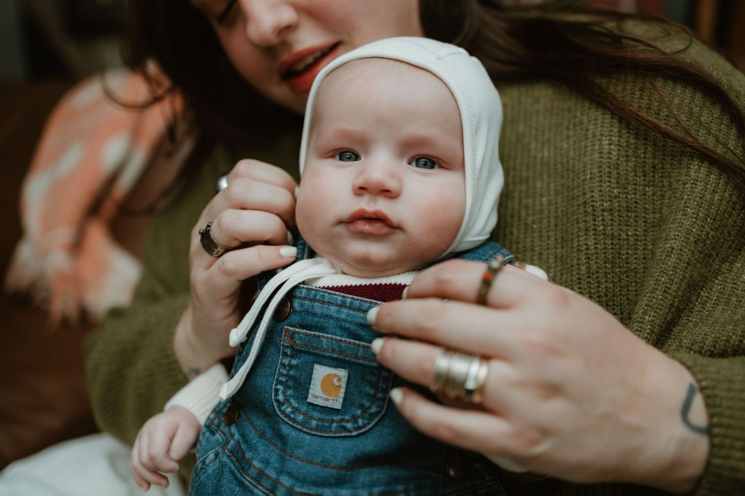 A mother in a green sweater adjusts a white bonnet on her baby wearing denim overalls.
