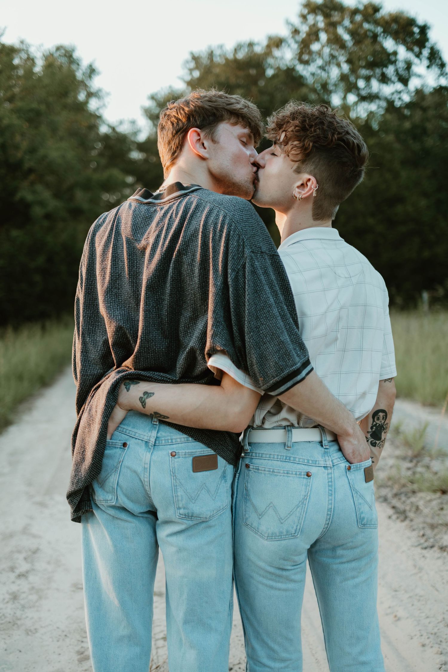Two people wearing jeans embrace and share a tender kiss on a dirt path surrounded by trees at dusk.