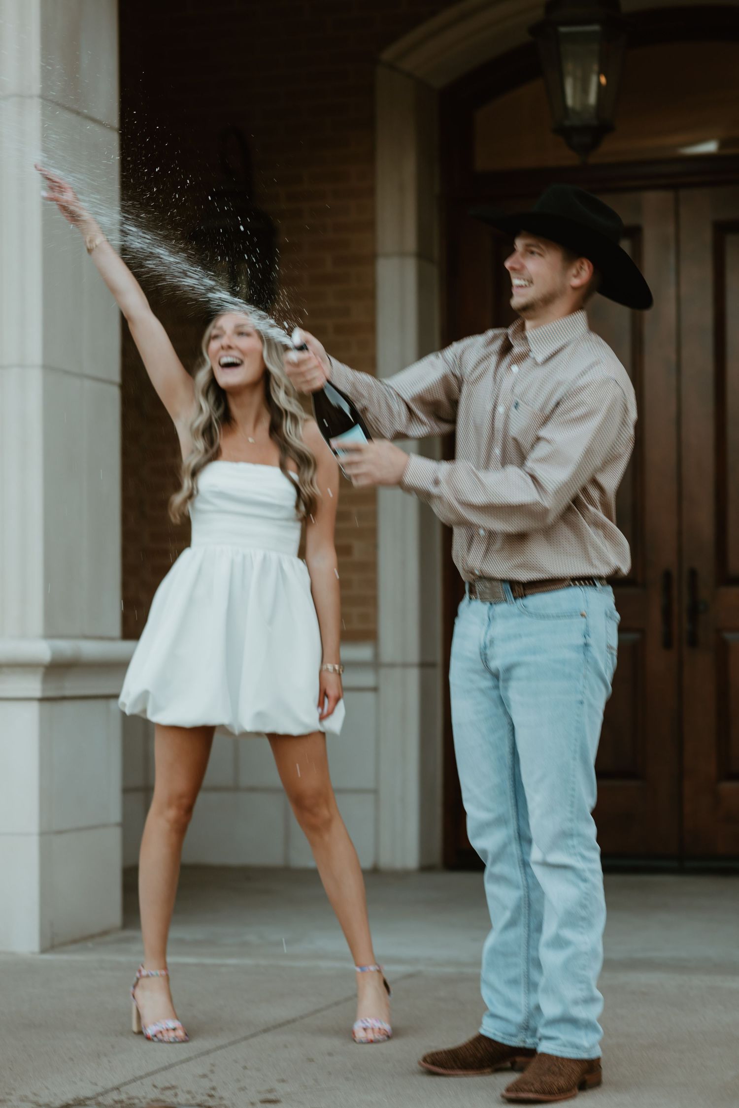 A couple celebrates with champagne spraying in the air, woman in white dress and man wearing cowboy hat and jeans.