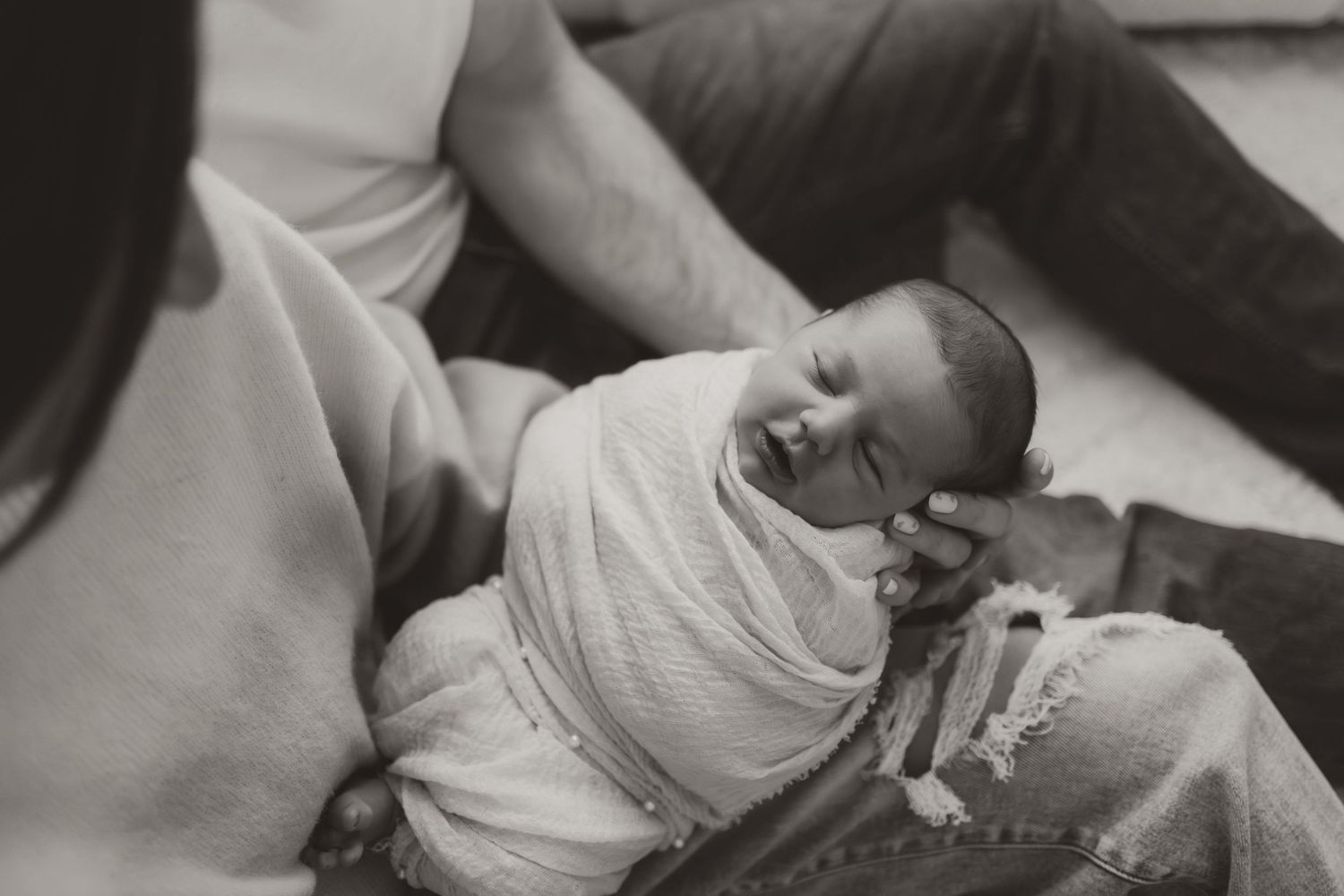 Black and white photo of a swaddled newborn baby smiling while cradled in parents arms.